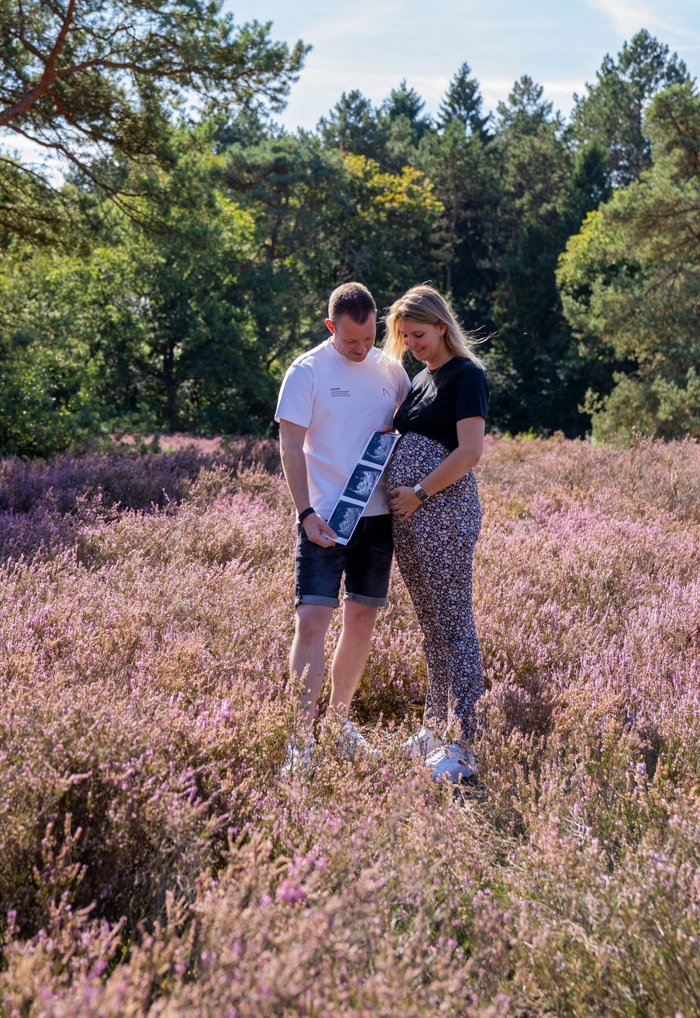 Een stel staat in een veld vol bloeiende paarse heide, met echofoto's in de hand, en kondigt in de buitenlucht hun zwangerschap aan.