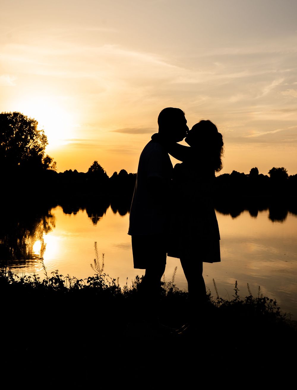 Romantisch silhouet van een stel dat elkaar kust bij een gouden zonsondergang die weerspiegeld wordt in het kalme water van een meer.
