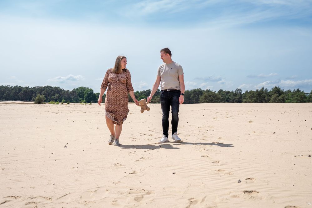 Een stel loopt hand in hand over zandduinen met dennenbomen en een blauwe lucht op de achtergrond.