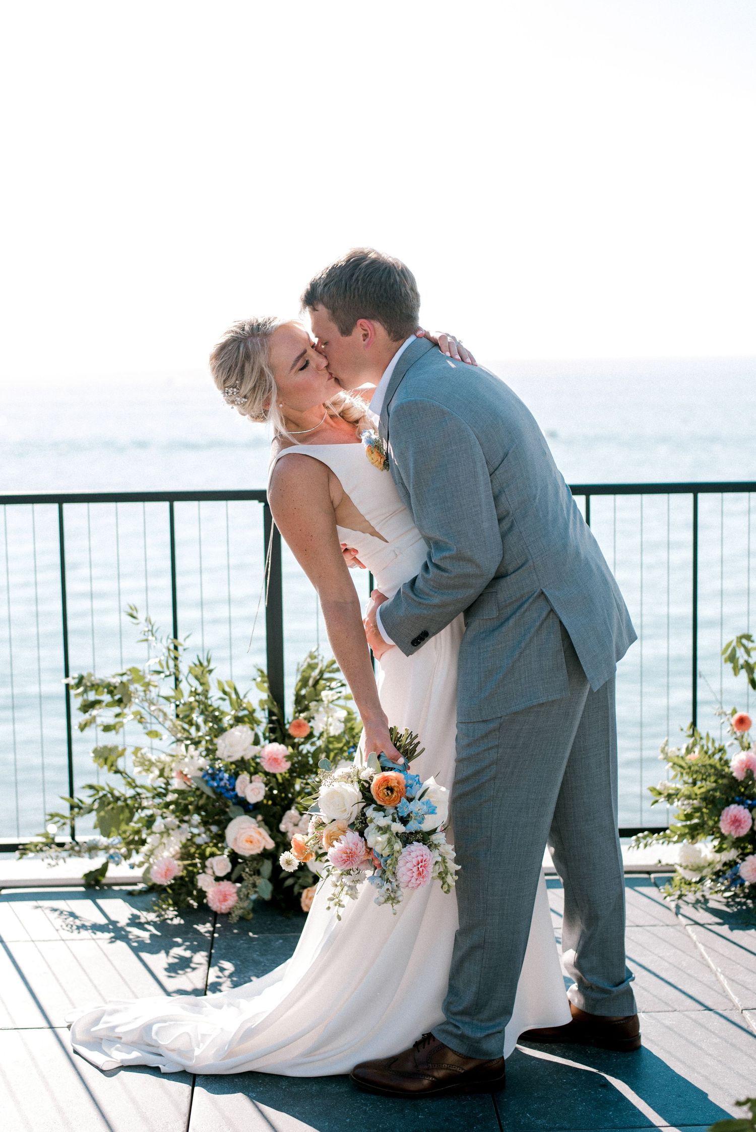 Bride and groom share first kiss at oceanfront wedding ceremony surrounded by colorful floral arrangements.