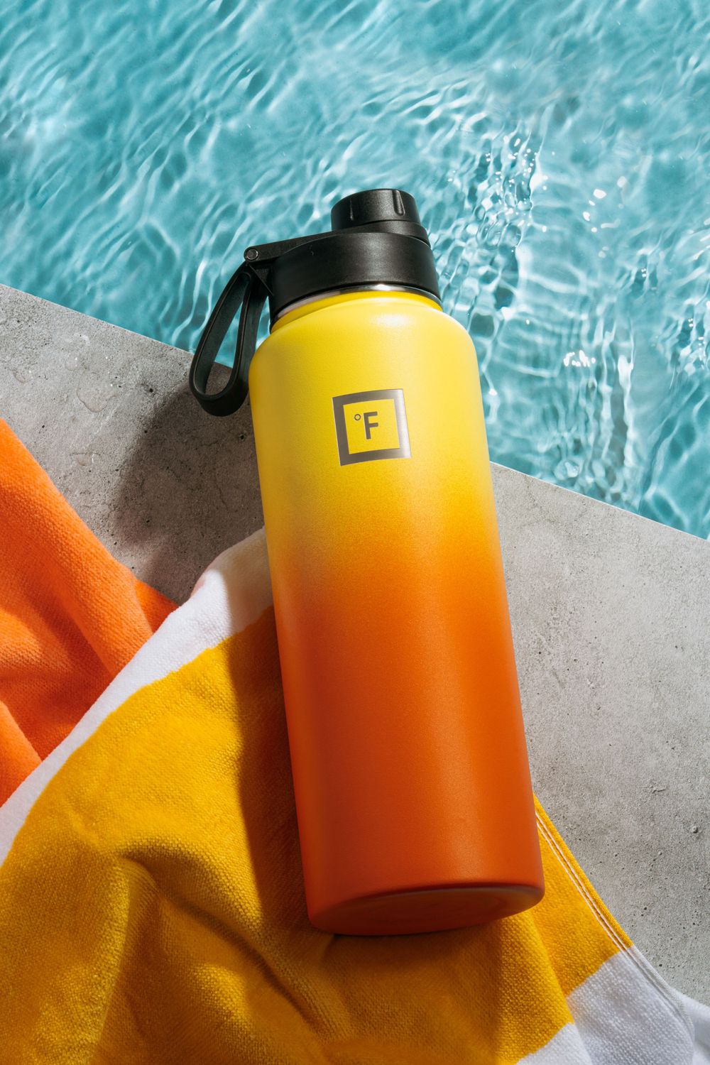 Yellow and orange gradient water bottle on towel by pool with turquoise water.