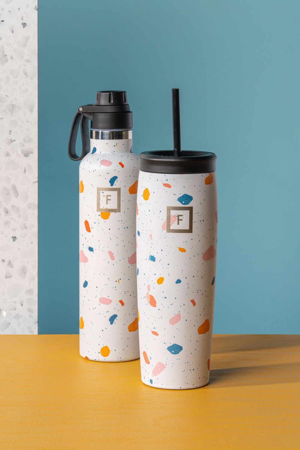 Two matching terrazzo patterned drink containers on yellow surface against blue wall.