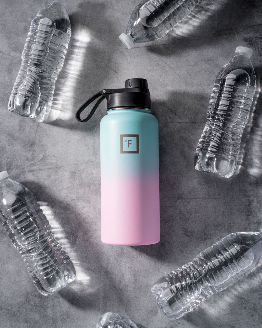 Reusable water bottle in pastel blue and pink surrounded by single-use plastic bottles on concrete surface.