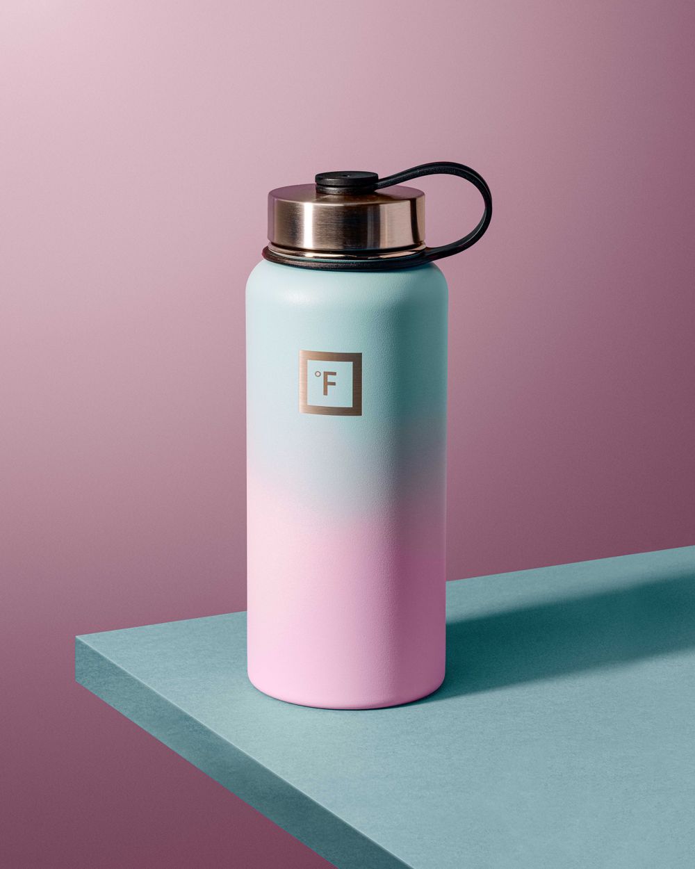 Gradient mint and pink water bottle with metallic cap on turquoise surface against pink background.