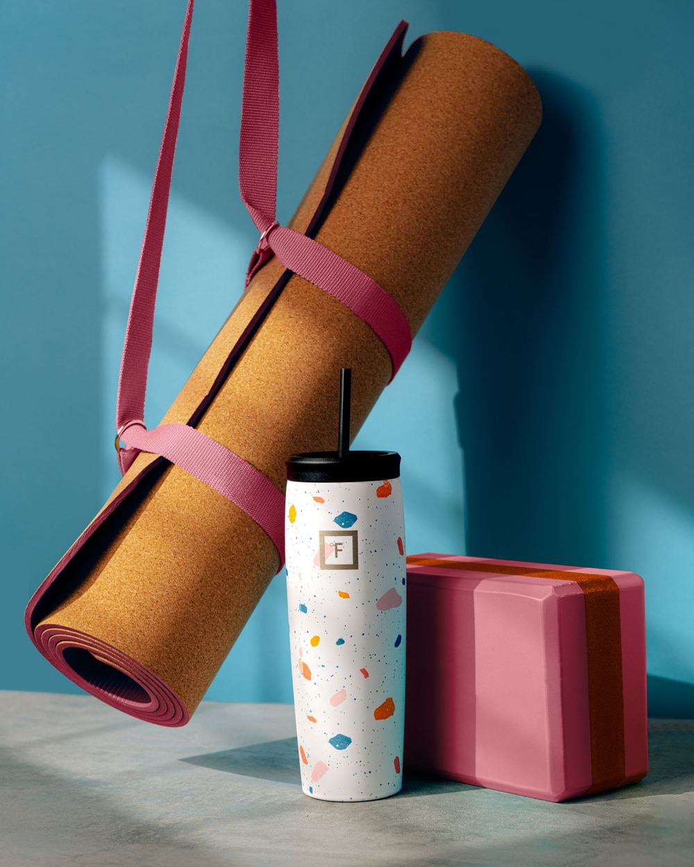Cork yoga mat with pink straps and patterned water bottle against blue background.