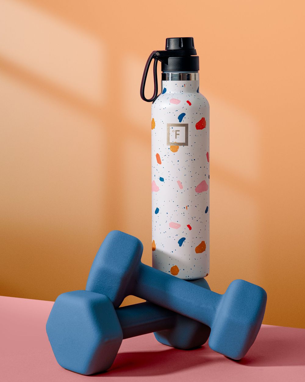 White water bottle with colorful terrazzo pattern next to blue dumbbells on pink and orange background.