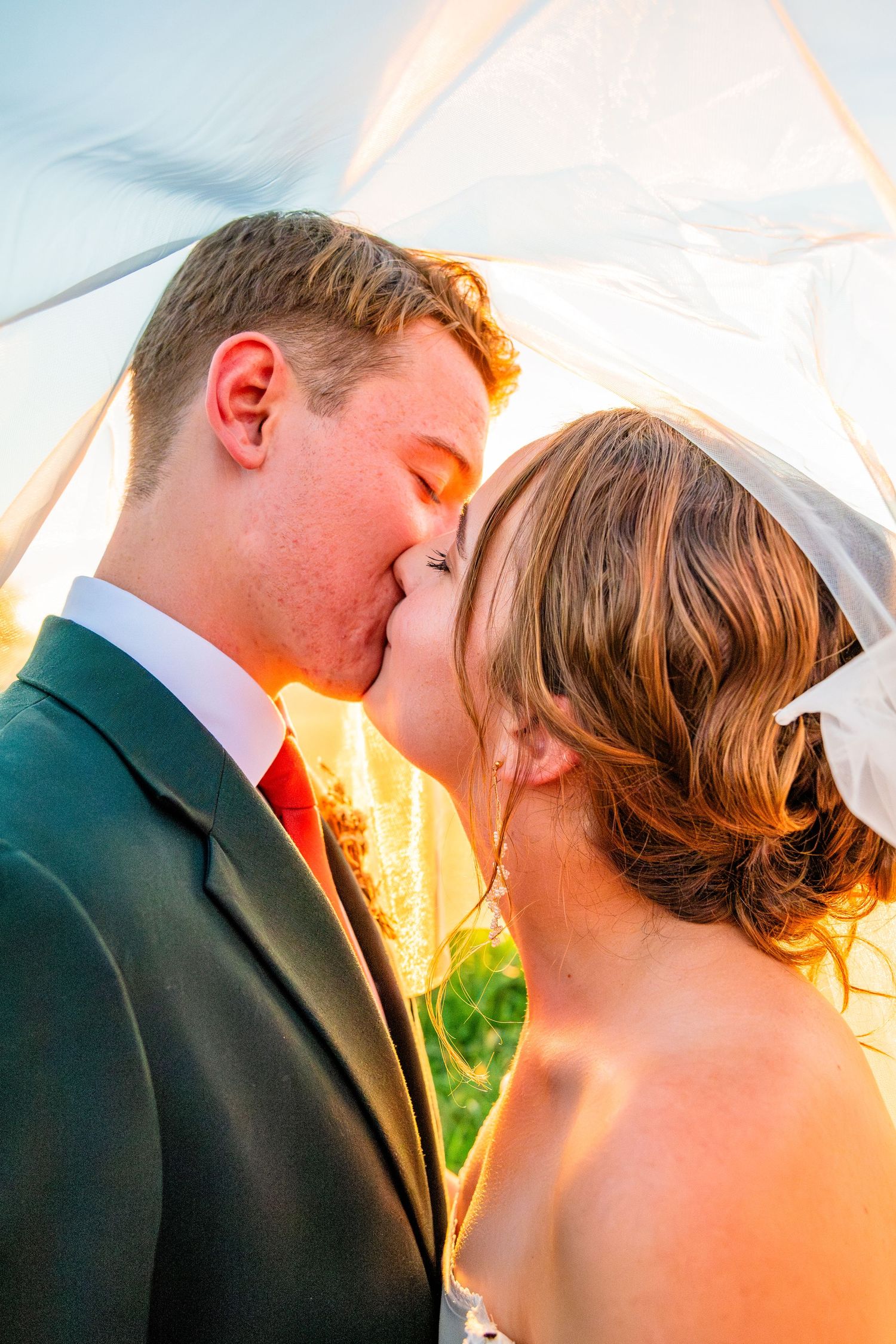 Bride and groom sharing a romantic kiss under a wedding veil with warm golden sunset light glowing behind them.
