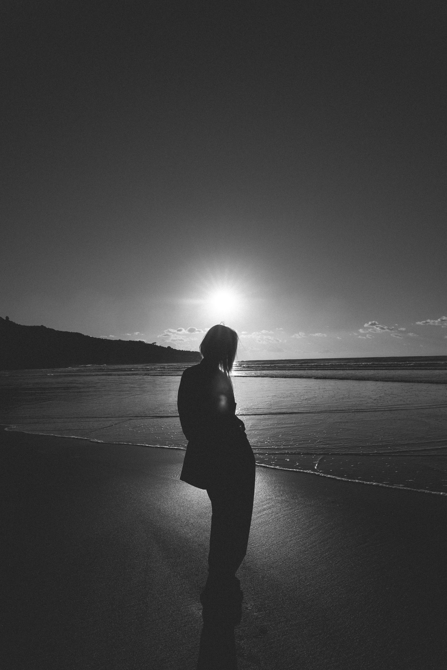 Black and white silhouette of a woman standing on a beach at sunset with sun glowing behind her in New Zealand.
