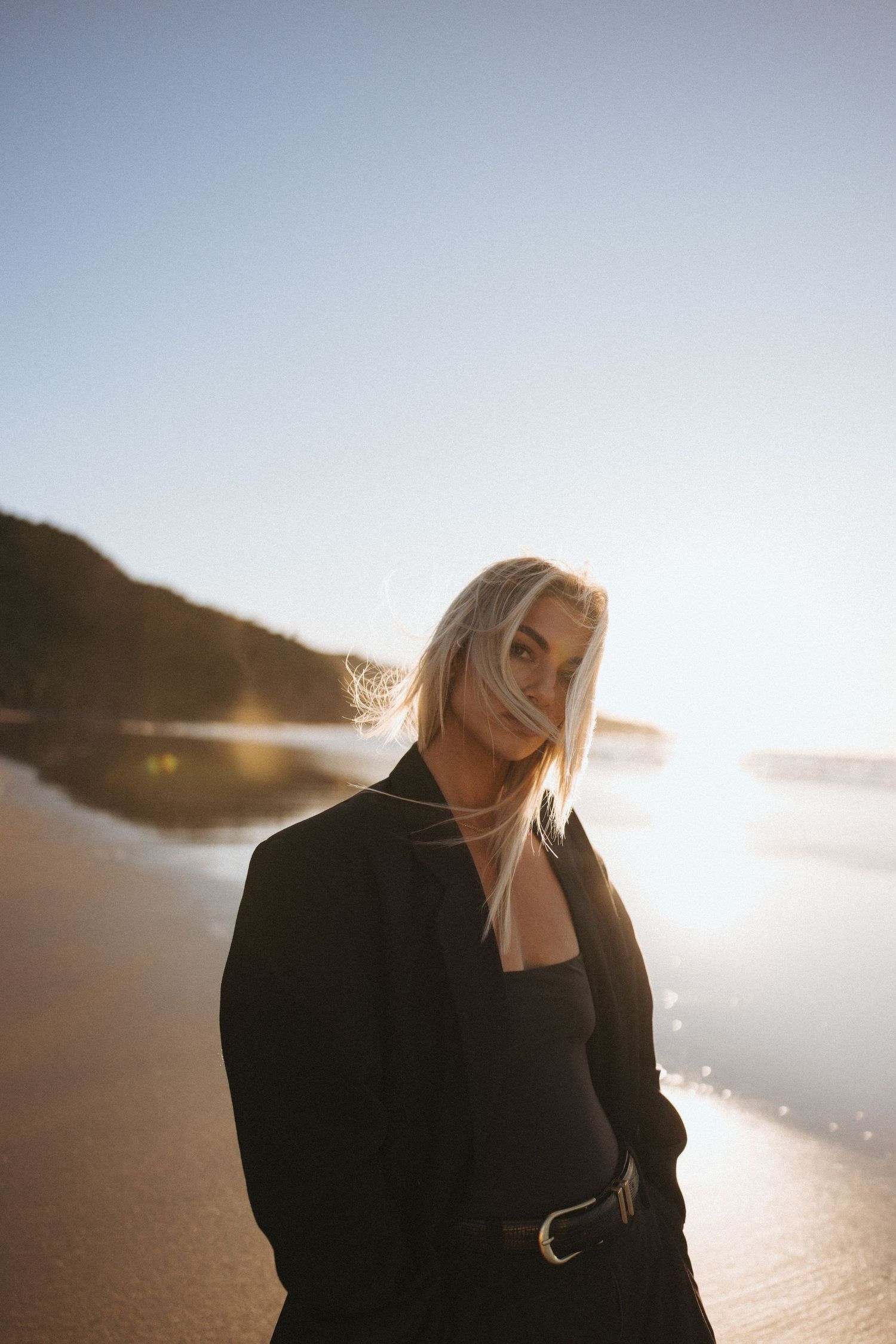 Blonde woman in black blazer standing on a sunlit beach with wind blowing her hair at golden hour in New Zealand.
