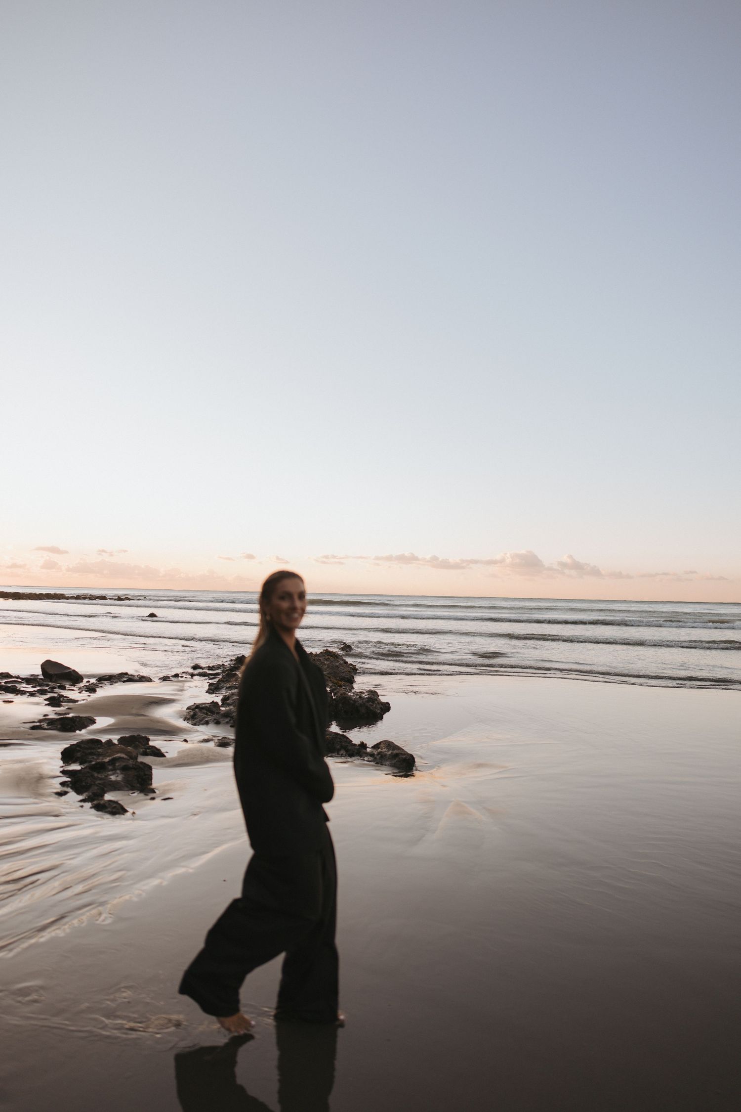 Smiling woman in black outfit walking barefoot on a reflective wet beach at twilight by rocky shore in New Zealand.