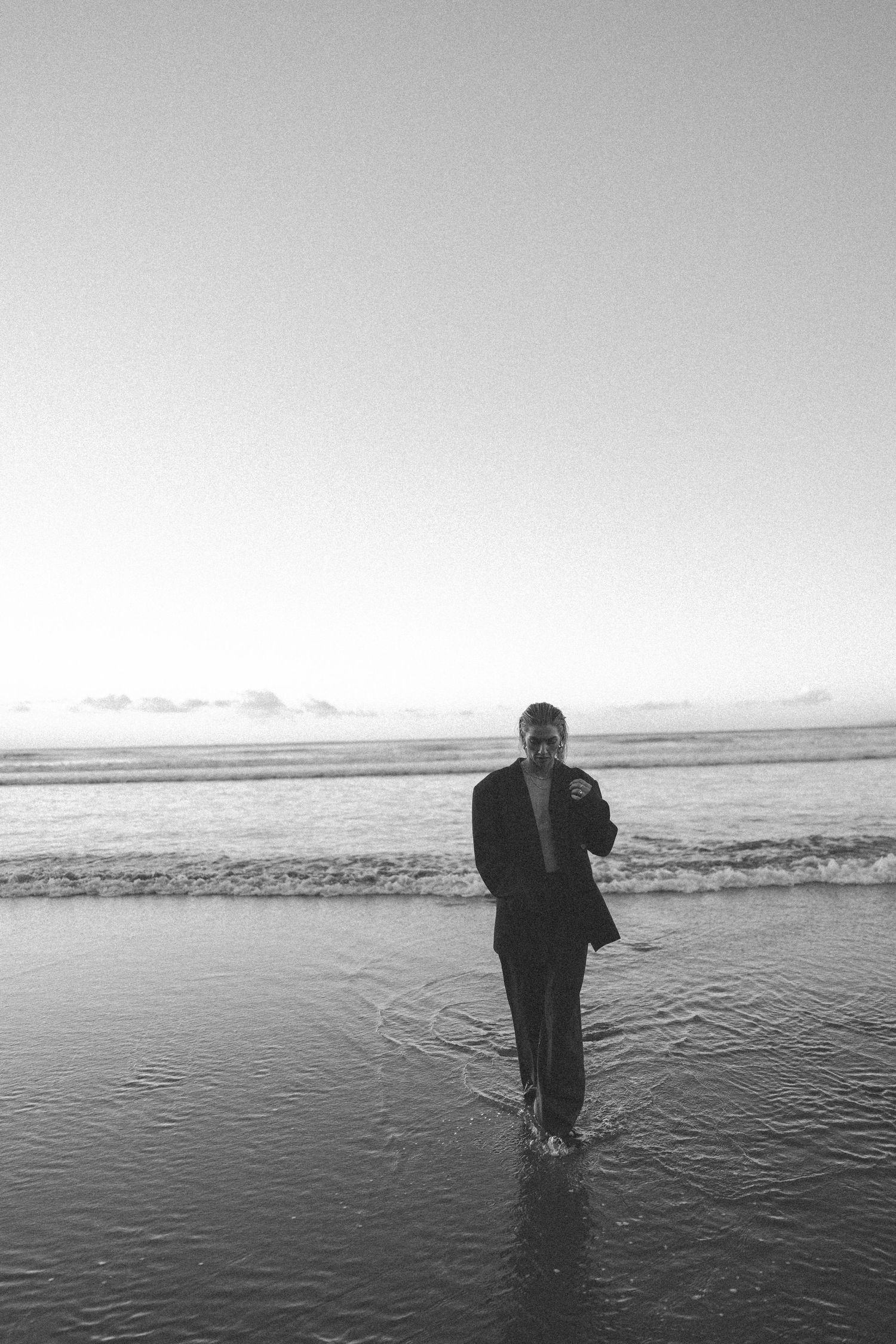 Black and white photo of a barefoot woman in a blazer standing in shallow ocean waves at sunset in New Zealand.