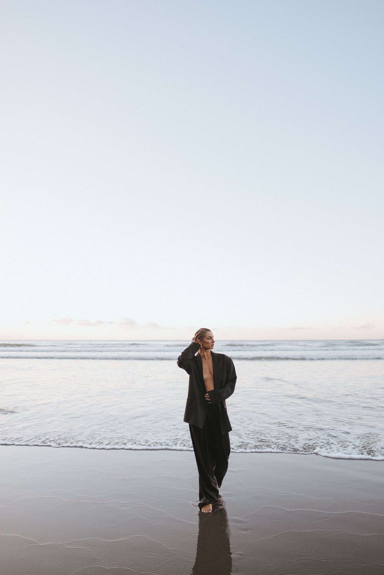 Woman in black blazer and pants standing barefoot on a serene beach at dusk with gentle ocean waves in New Zealand.