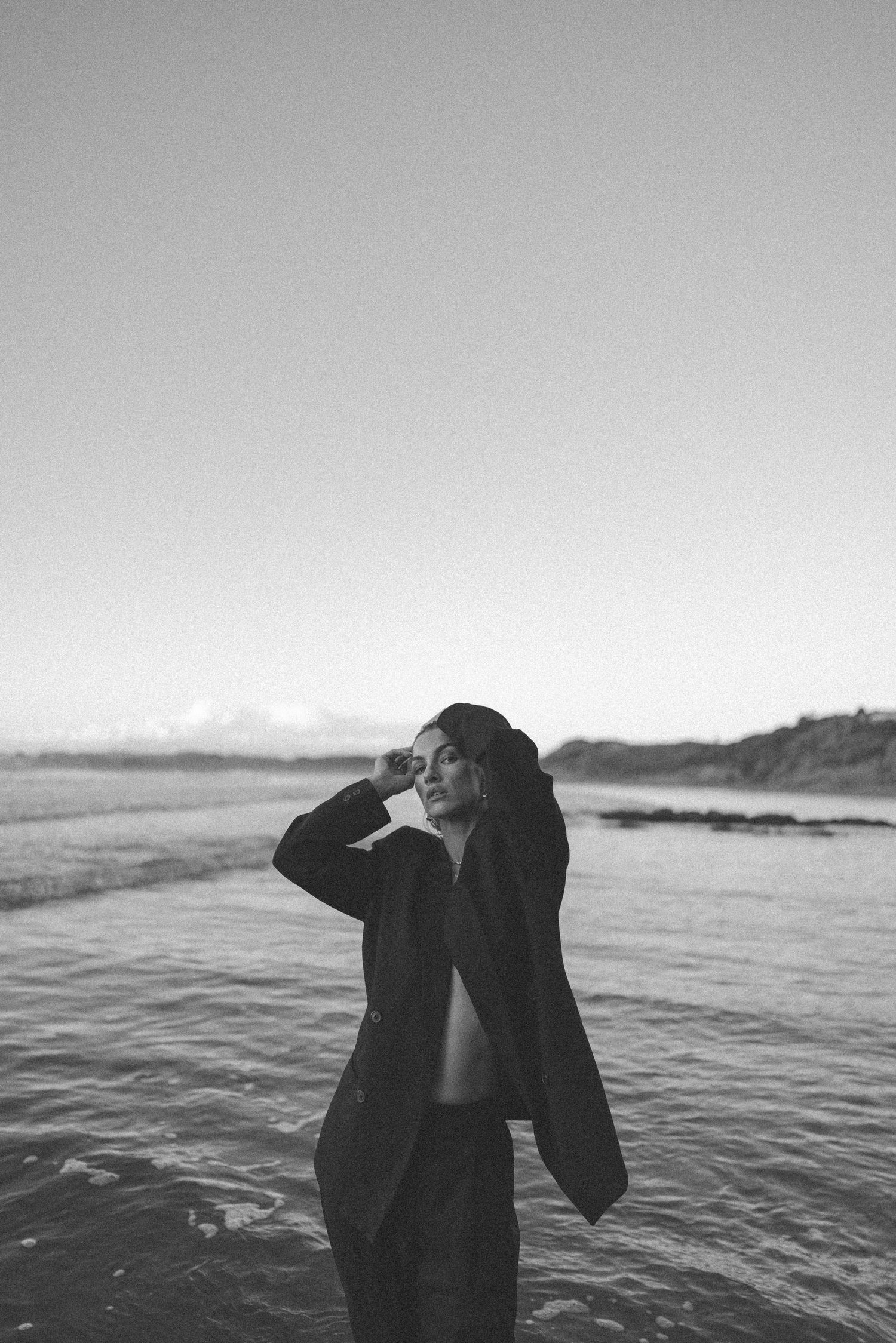 Black and white portrait of a woman in an oversized blazer posing in ocean water at dusk on beach in New Zealand.