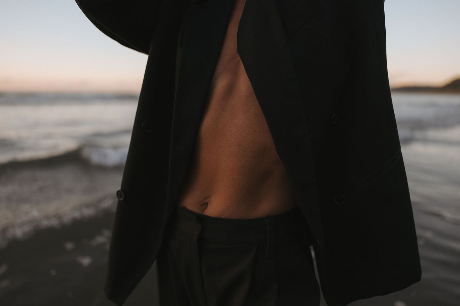 Close-up of a person wearing an open black blazer revealing bare torso with a small tattoo at sunset beach in New Zealand.