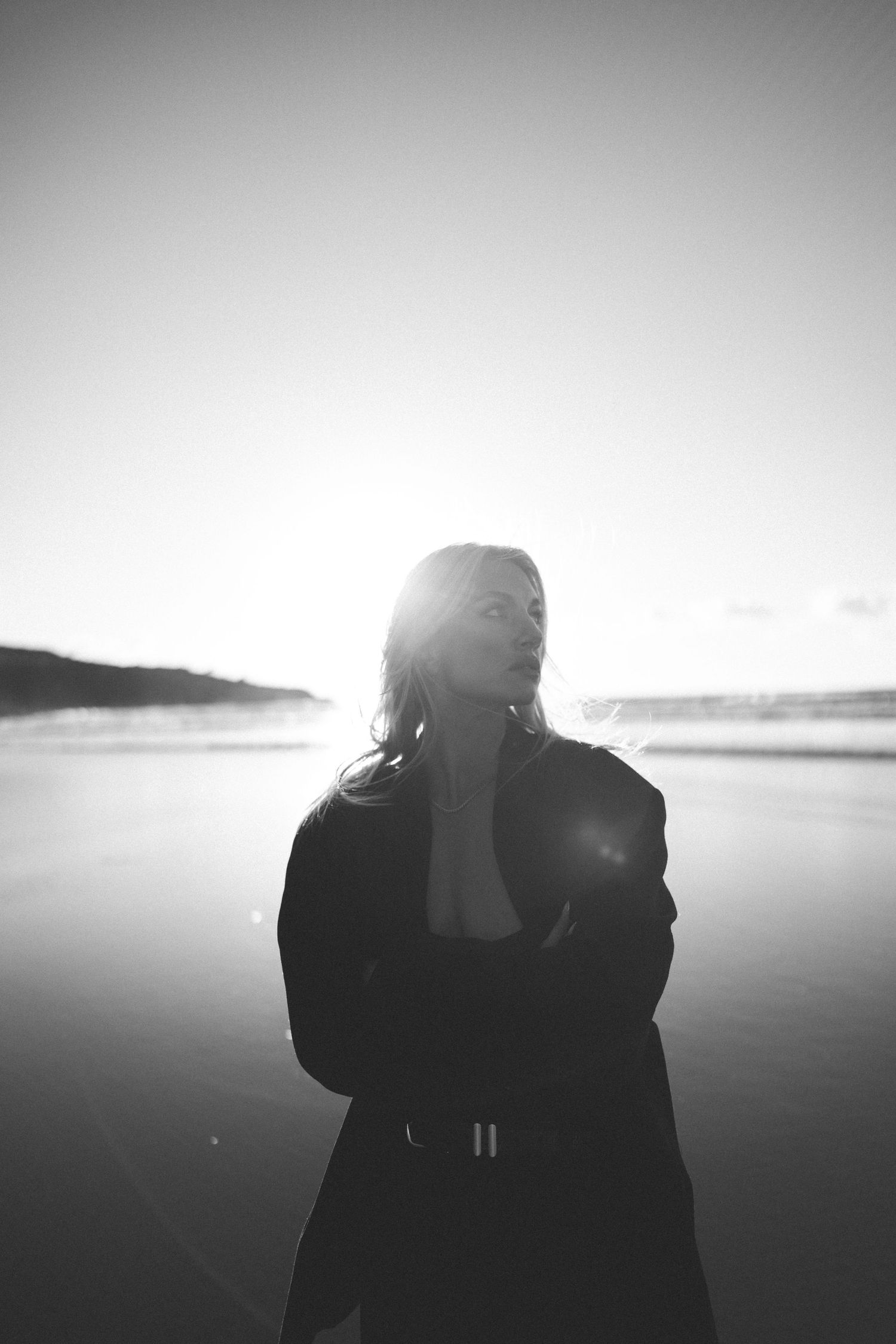 Black and white portrait of a woman standing on a reflective beach with bright sun flare behind her in New Zealand.