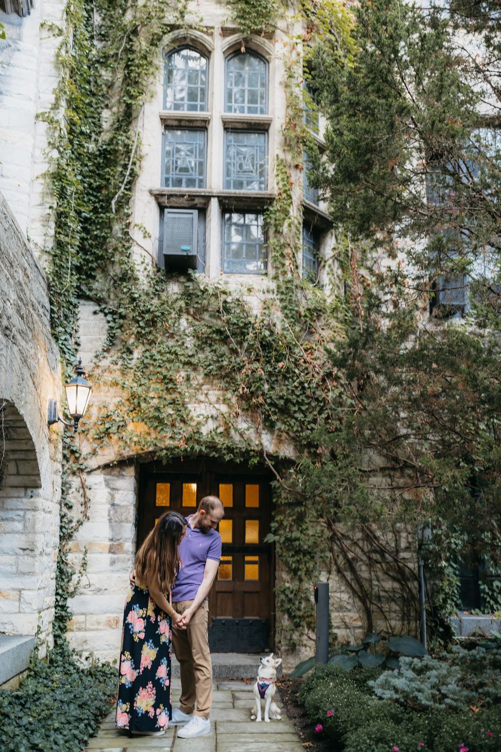 Couple standing in lush garden of Northwestern University grounds during engagement session accompanied by a small Corgi dog