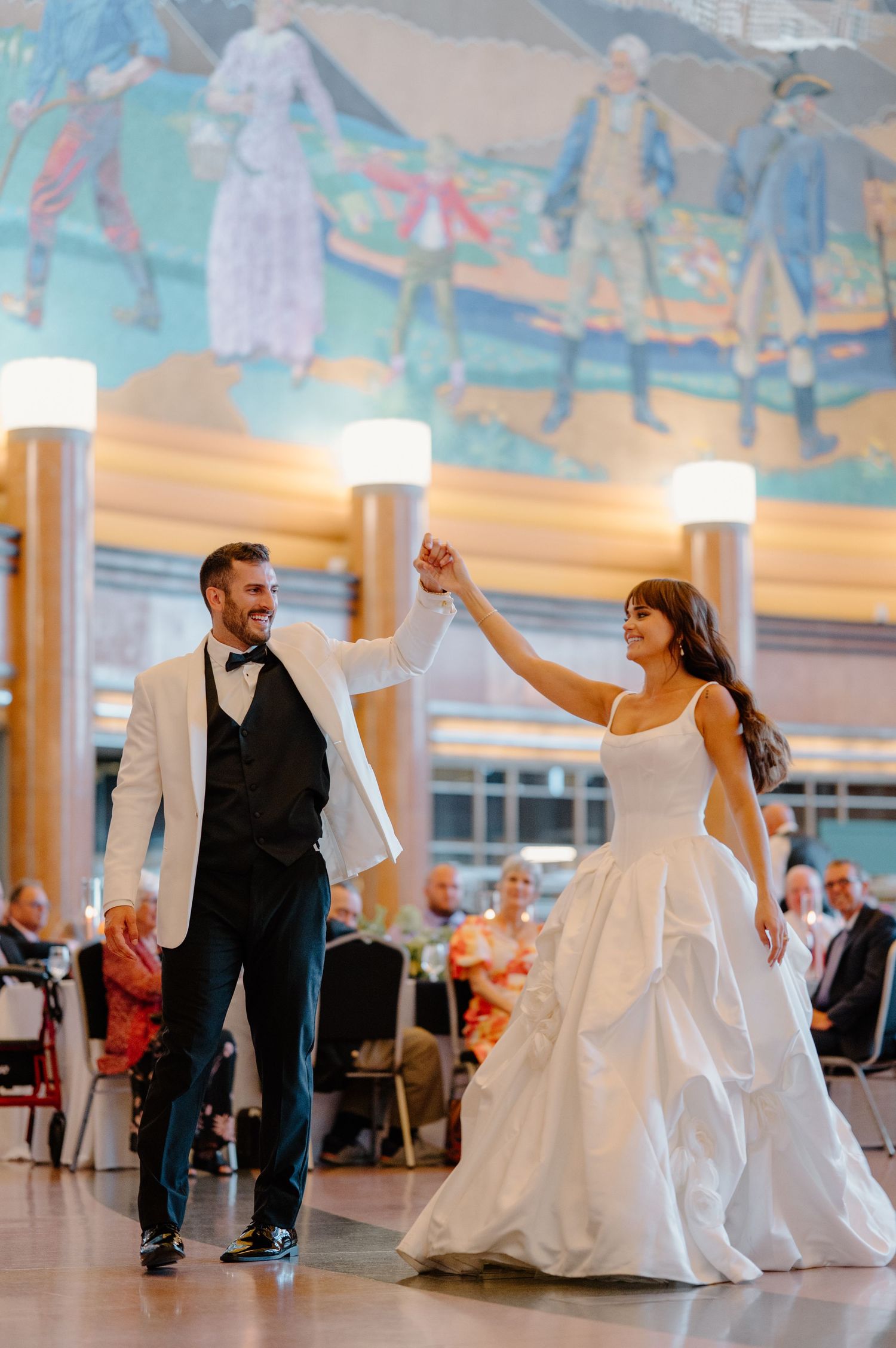 Bride and groom share first dance in elegant venue with colorful mural backdrop, groom in white tuxedo jacket.