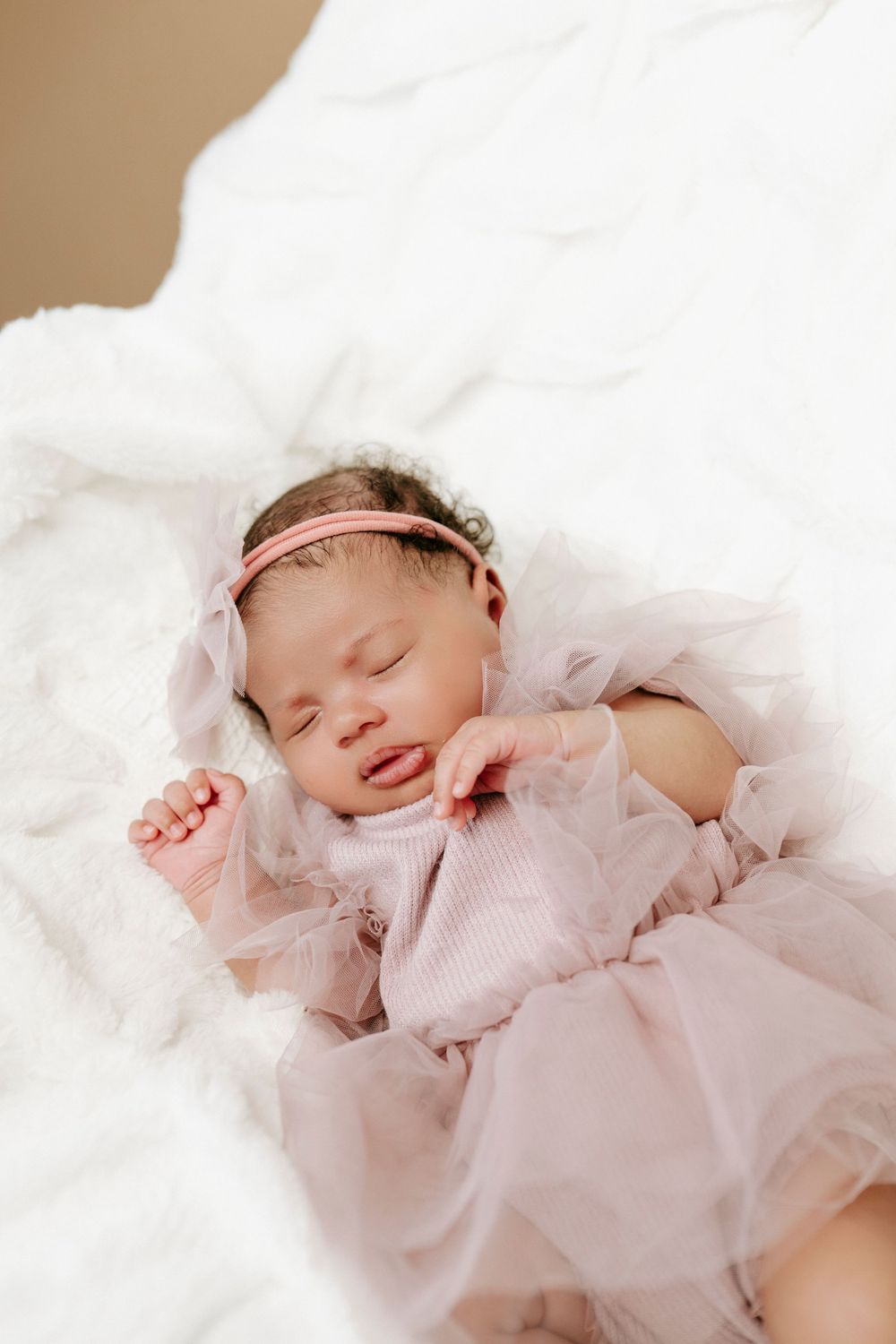 Sleeping newborn baby girl in pink tulle dress and headband on white fluffy blanket.