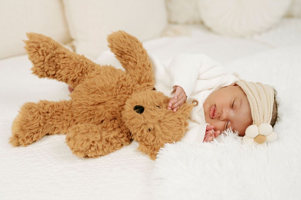 Newborn baby sleeping peacefully in white outfit and hat beside a fluffy brown teddy bear on a soft white blanket.