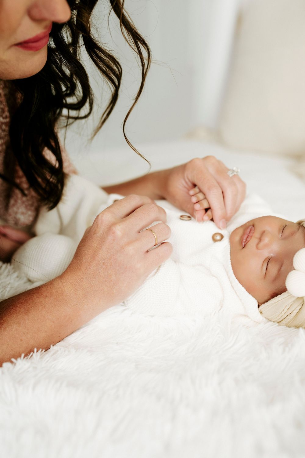 A mother with dark wavy hair tenderly holds her sleeping newborn baby dressed in a white outfit on a fluffy white blanket.