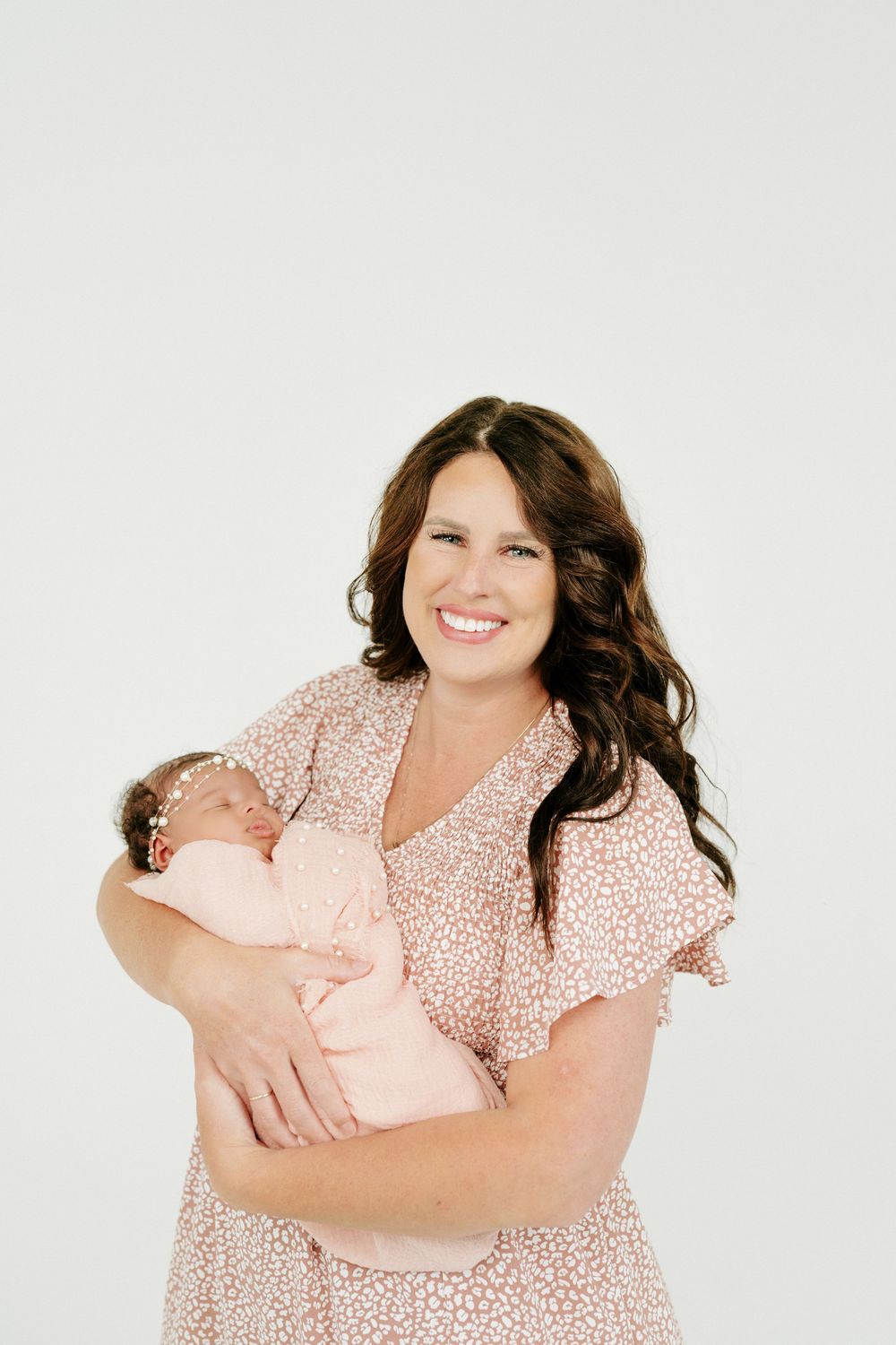 Smiling brunette woman in pink floral dress holding a swaddled newborn baby girl with pink headband.