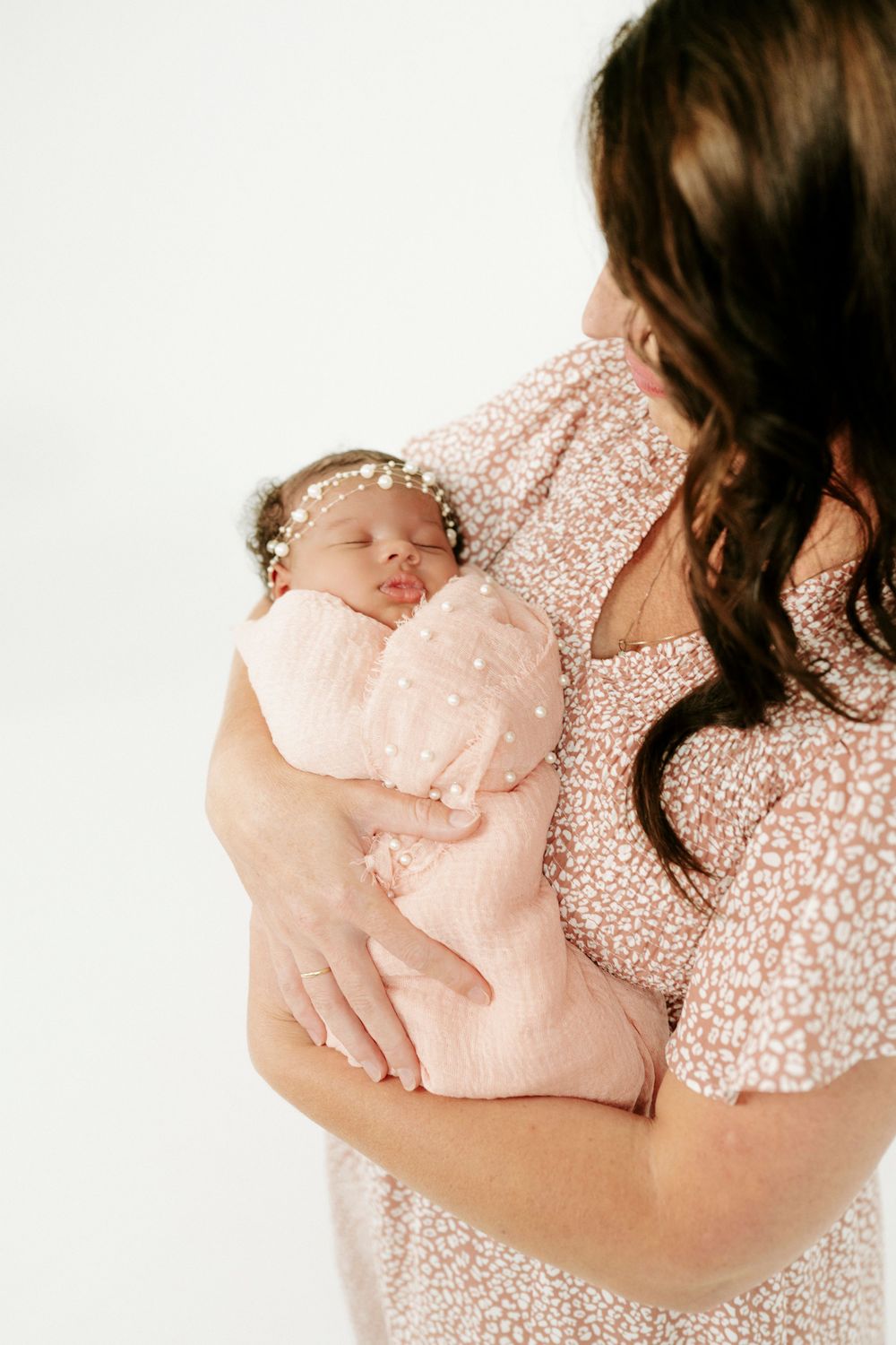 Mother in pink floral dress cradling sleeping newborn baby wrapped in pink swaddle.
