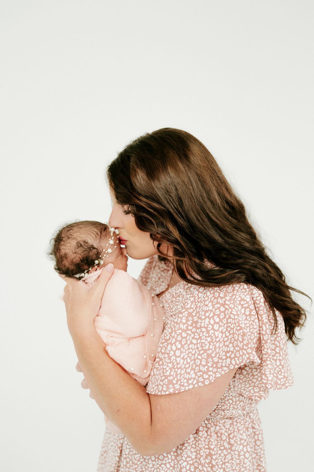 Brunette mother in pink dress gently kissing her sleeping newborn baby girl.