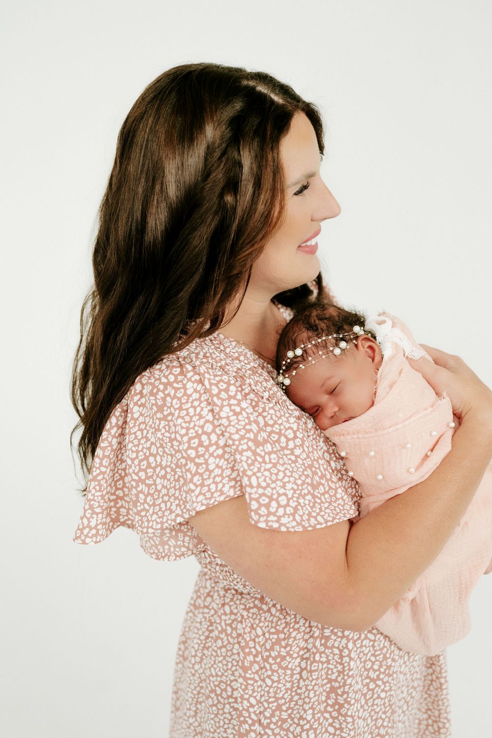 Smiling brunette mother holding sleeping newborn girl in pink swaddle with pearl headband.