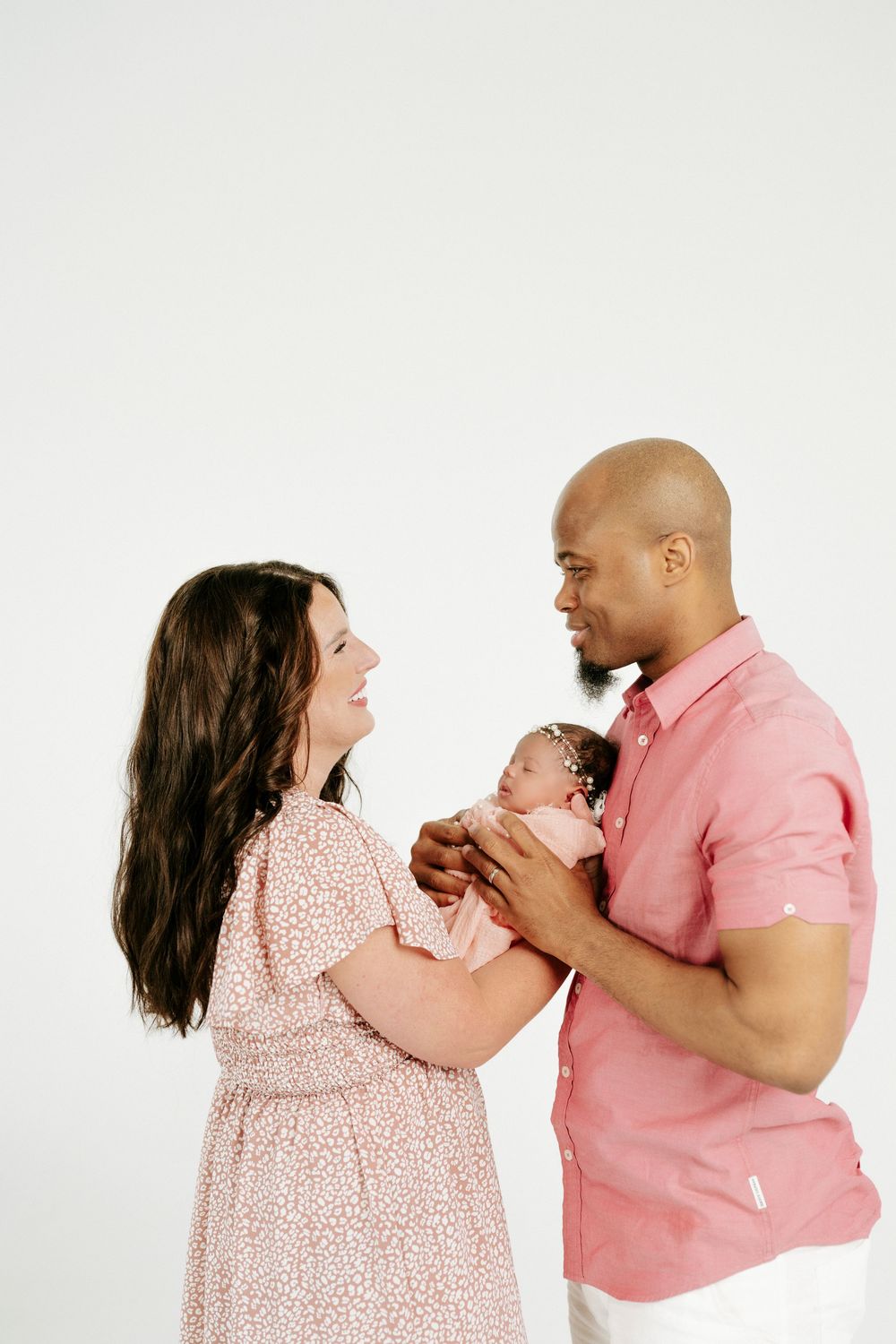A couple in pink outfits lovingly hold their newborn baby together against a white background.