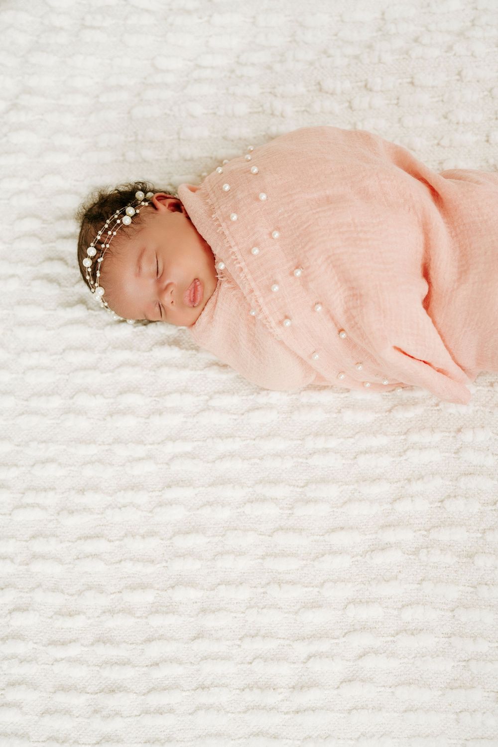 Sleeping newborn baby swaddled in pink wrap with pearl headband on white knit blanket.