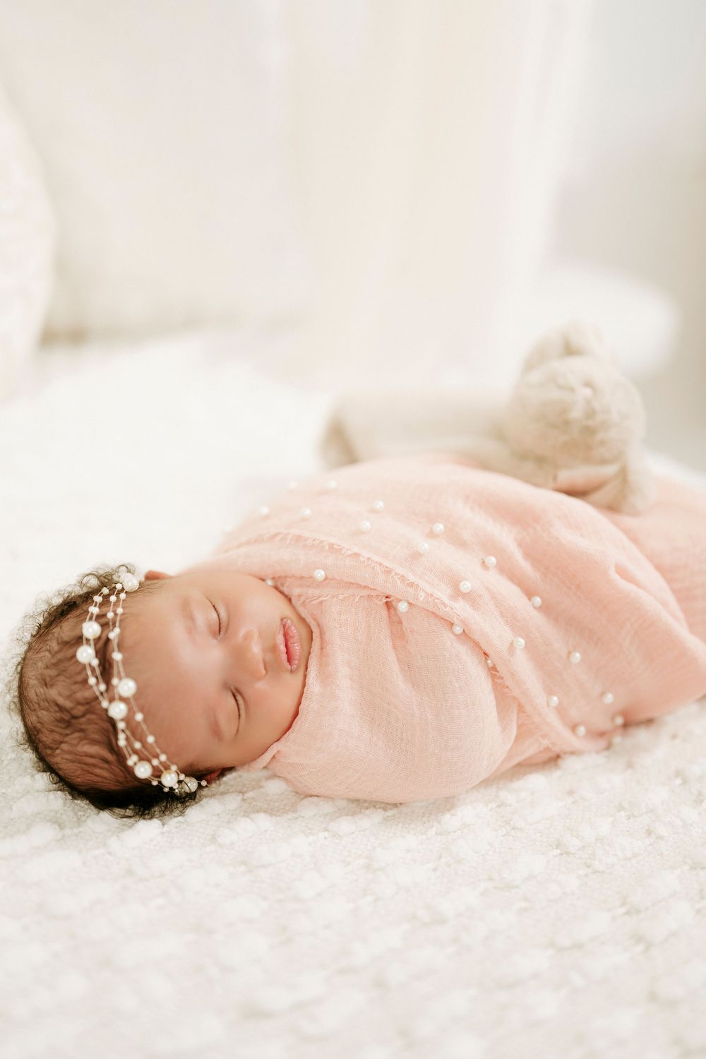 Peaceful newborn baby in pink swaddle with pearl headband sleeping on white fluffy blanket.