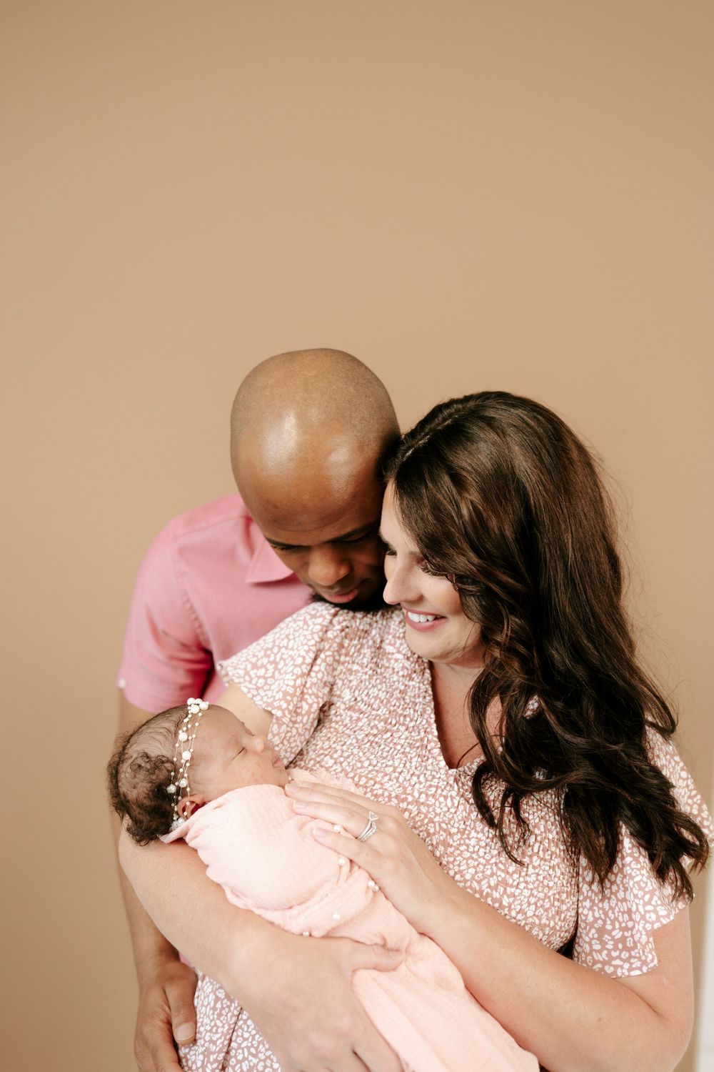 Happy couple smiling at their newborn baby wrapped in pink, studio portrait on beige background.