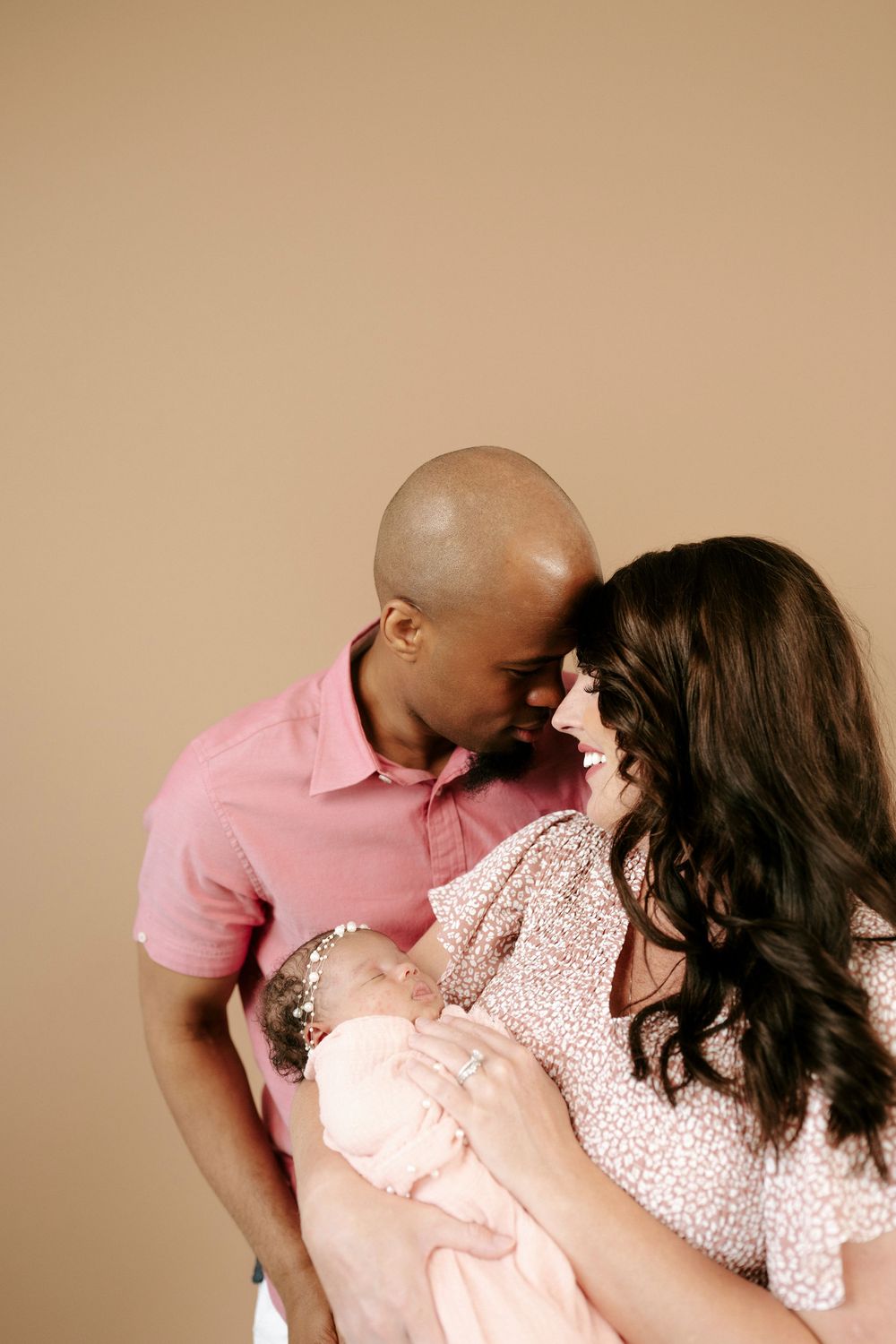 Happy interracial couple smiling at each other while cradling their newborn baby in a warm studio setting.