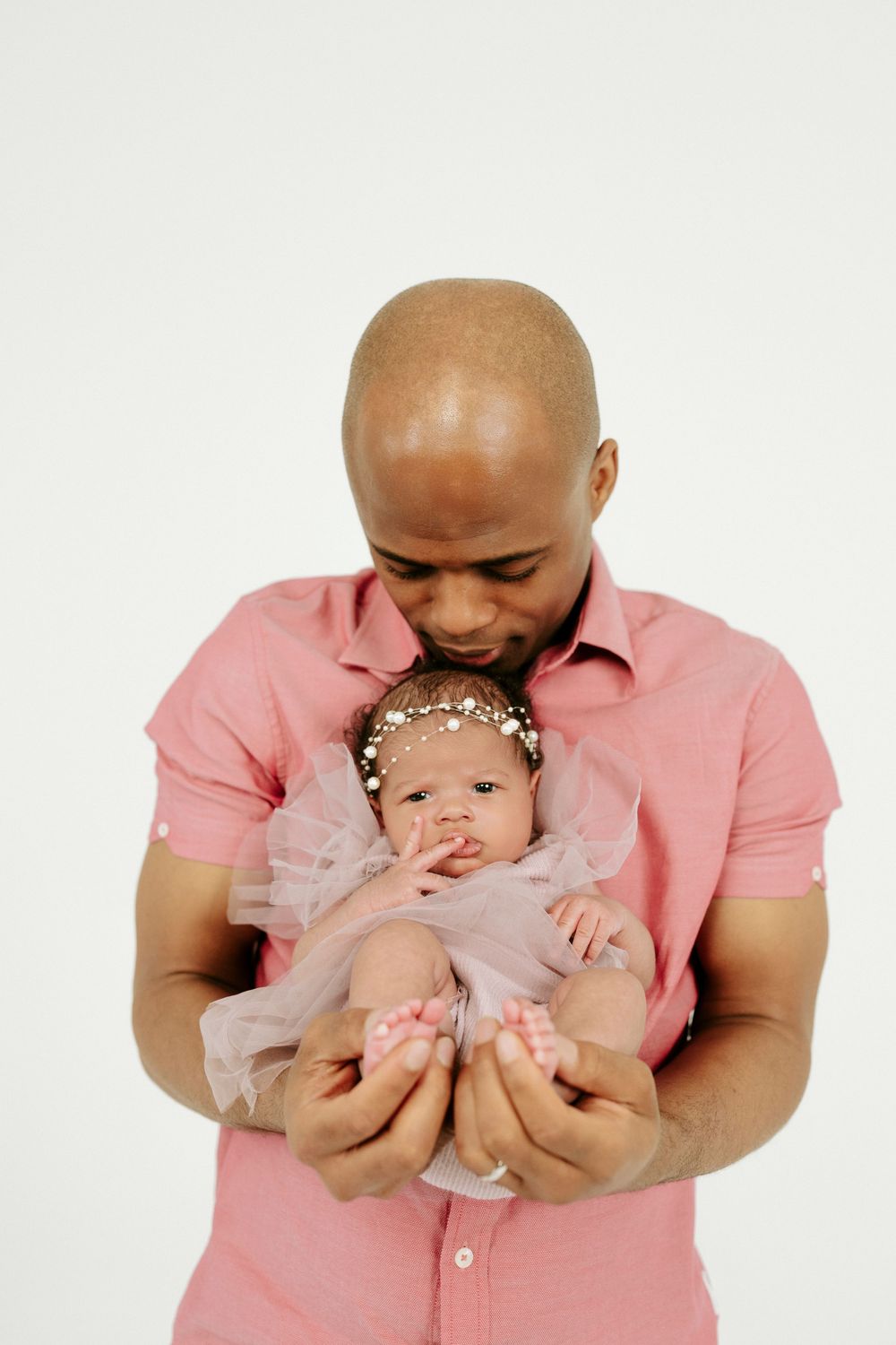 Bald father in pink shirt tenderly kisses newborn baby girl in tutu dress and pearl headband against white background.