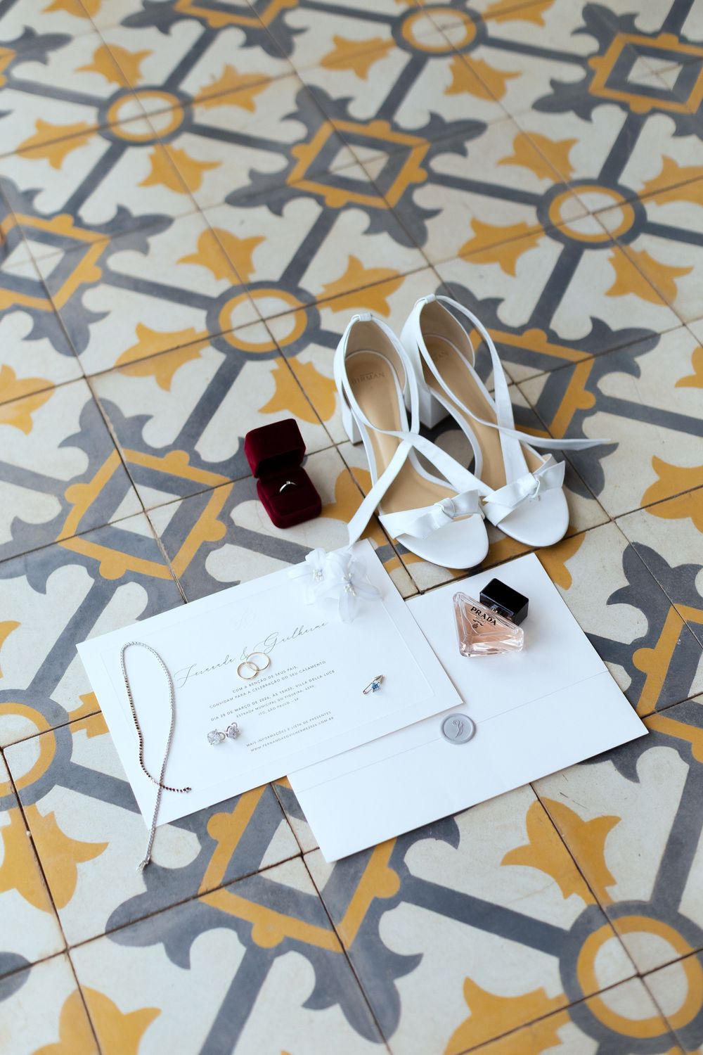 White bridal heels, wedding invitation, perfume, ring box, and earrings arranged on decorative yellow and gray tile floor.