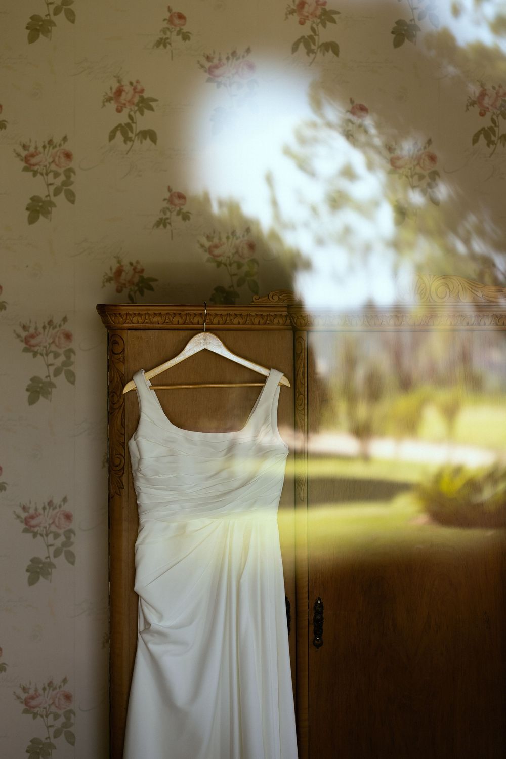 White wedding dress hanging on wooden armoire with floral wallpaper and garden reflection in window.
