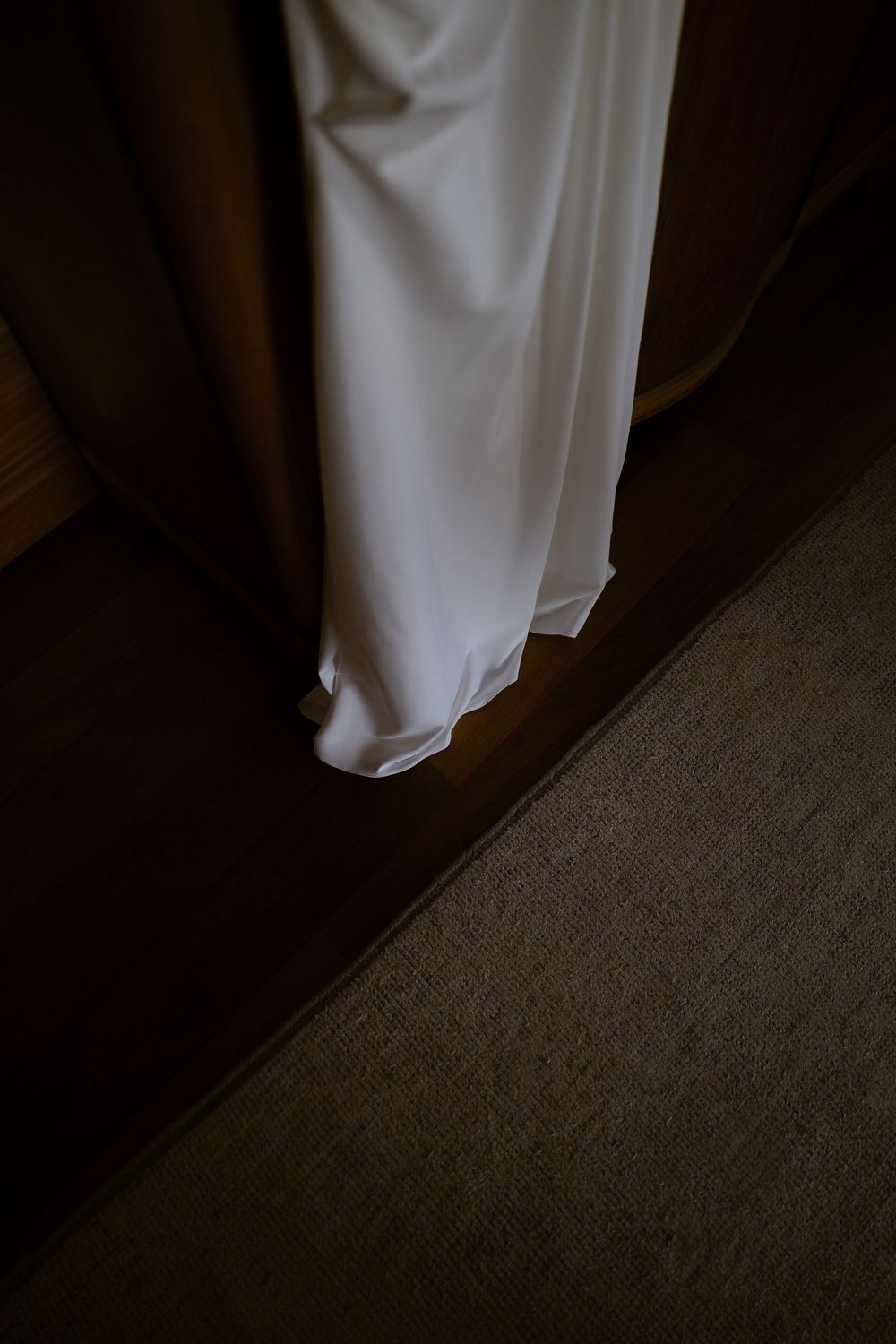 White wedding dress hanging in a dark wooden closet, cascading to the floor in soft elegant folds.