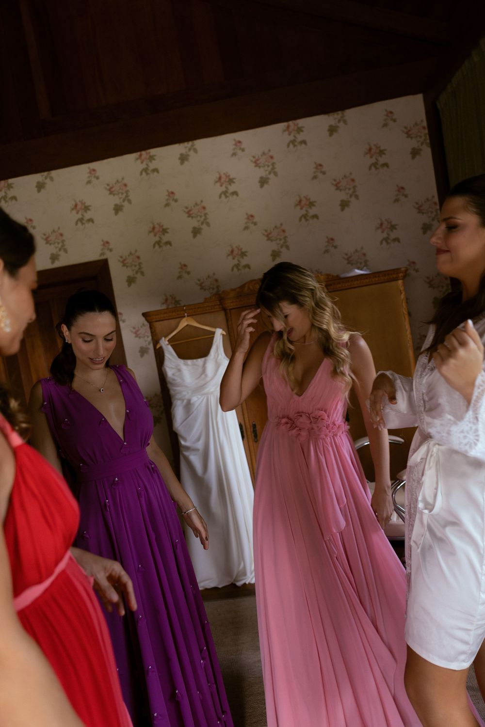 Bridesmaids in colorful gowns getting ready in a floral-wallpapered room with a white wedding dress hanging in background.
