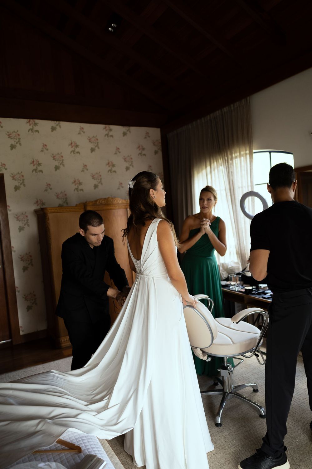 Bride in elegant white wedding gown being assisted by stylists in a warmly lit bridal preparation room.