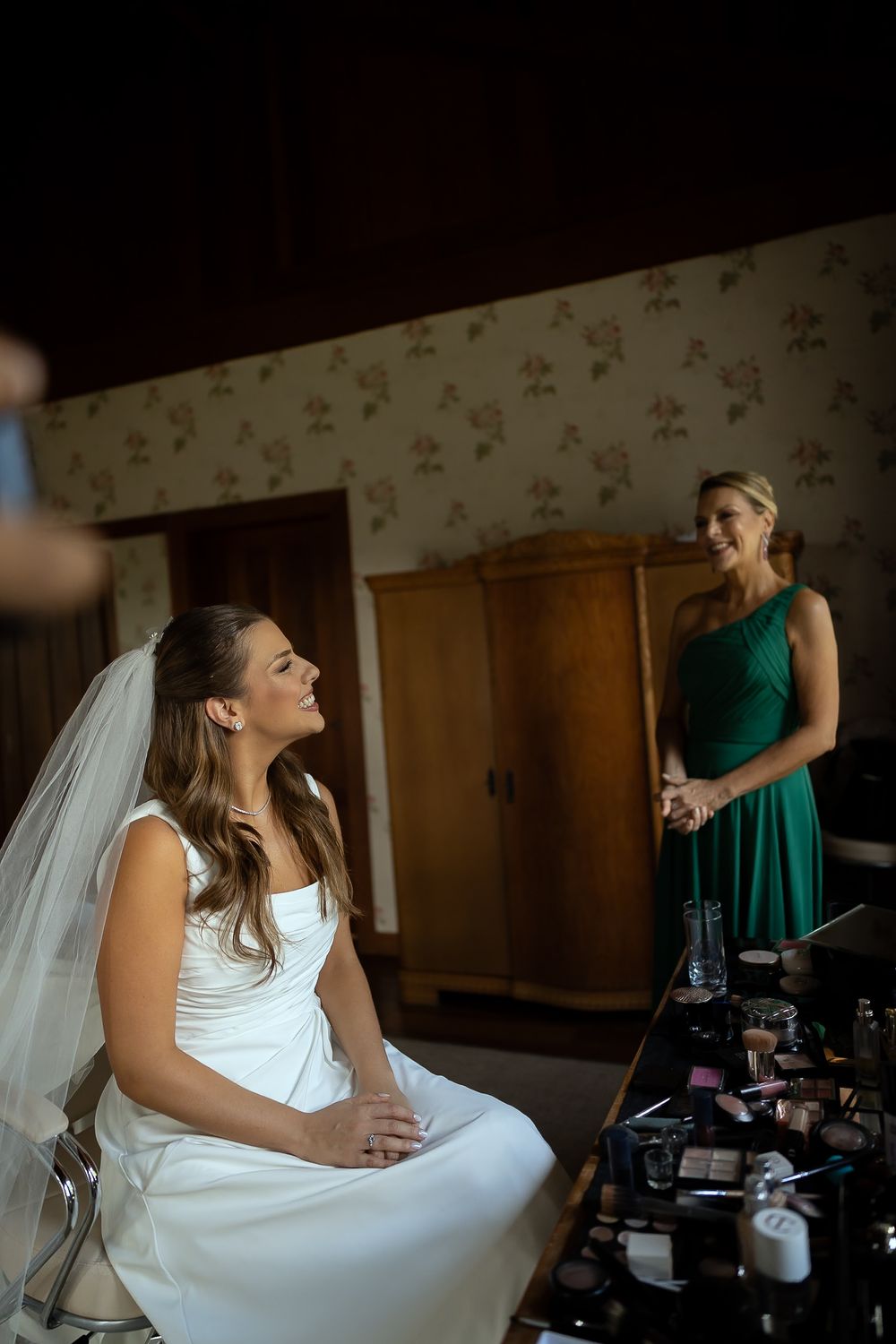 Bride in white gown and veil sits smiling while bridesmaid in green dress stands nearby in a decorated room.