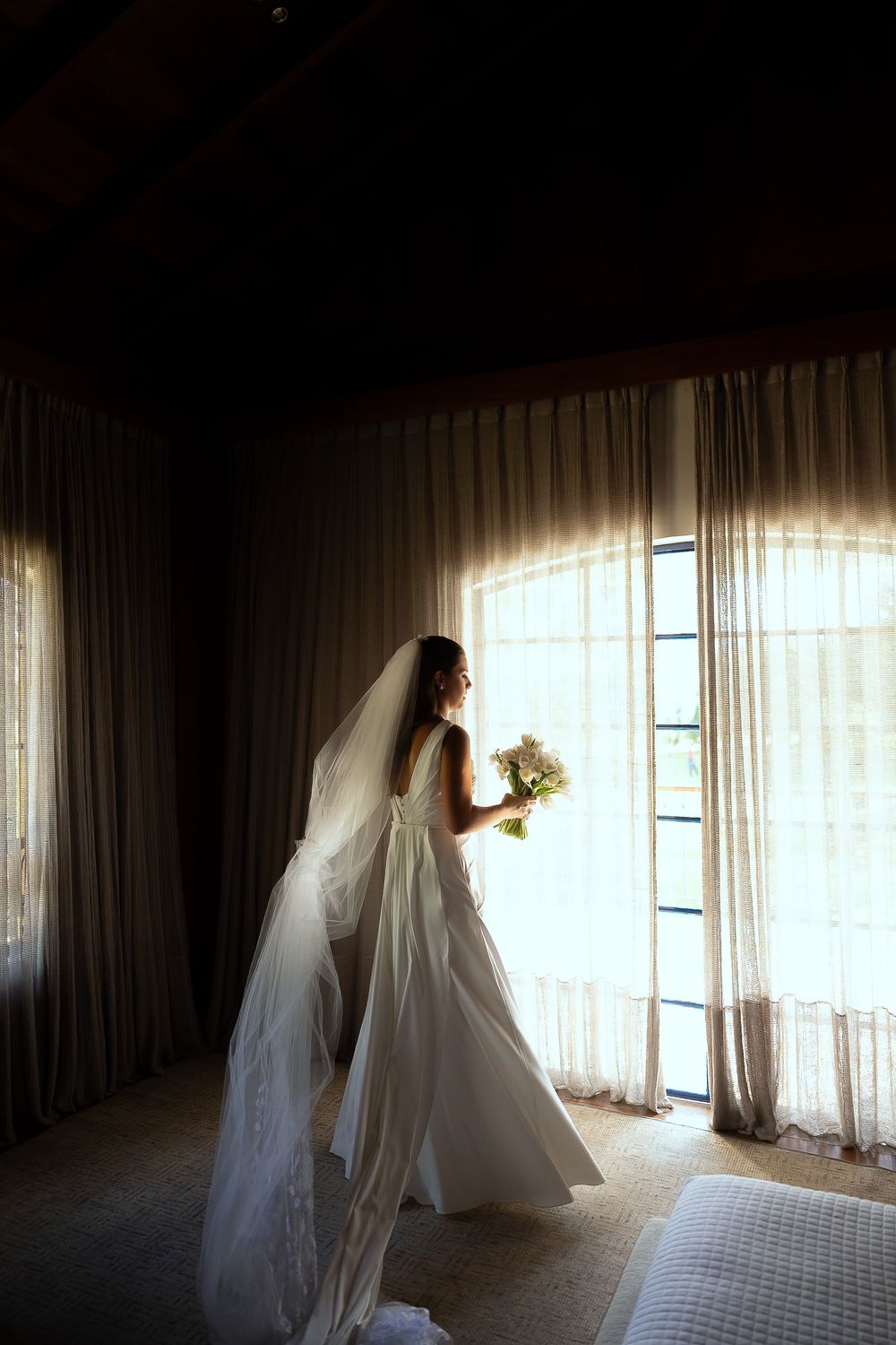 Bride in white gown and veil holding bouquet stands by sunlit curtained window in elegant dark room.