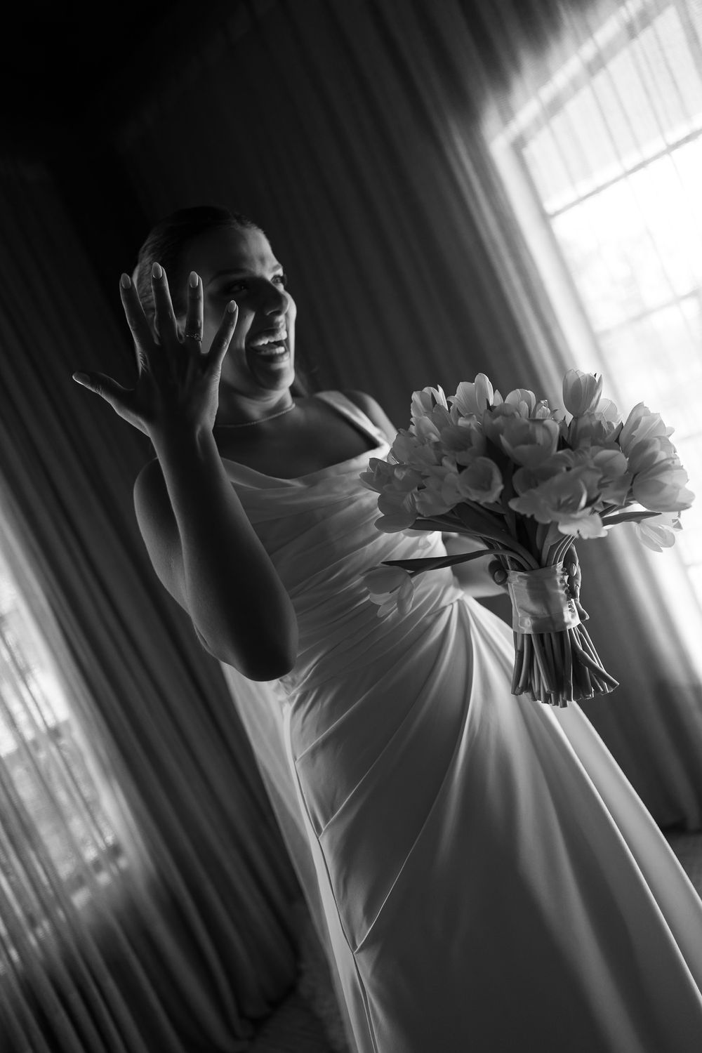 Black and white photo of a bride in a white gown holding a flower bouquet, smiling and raising her hand.