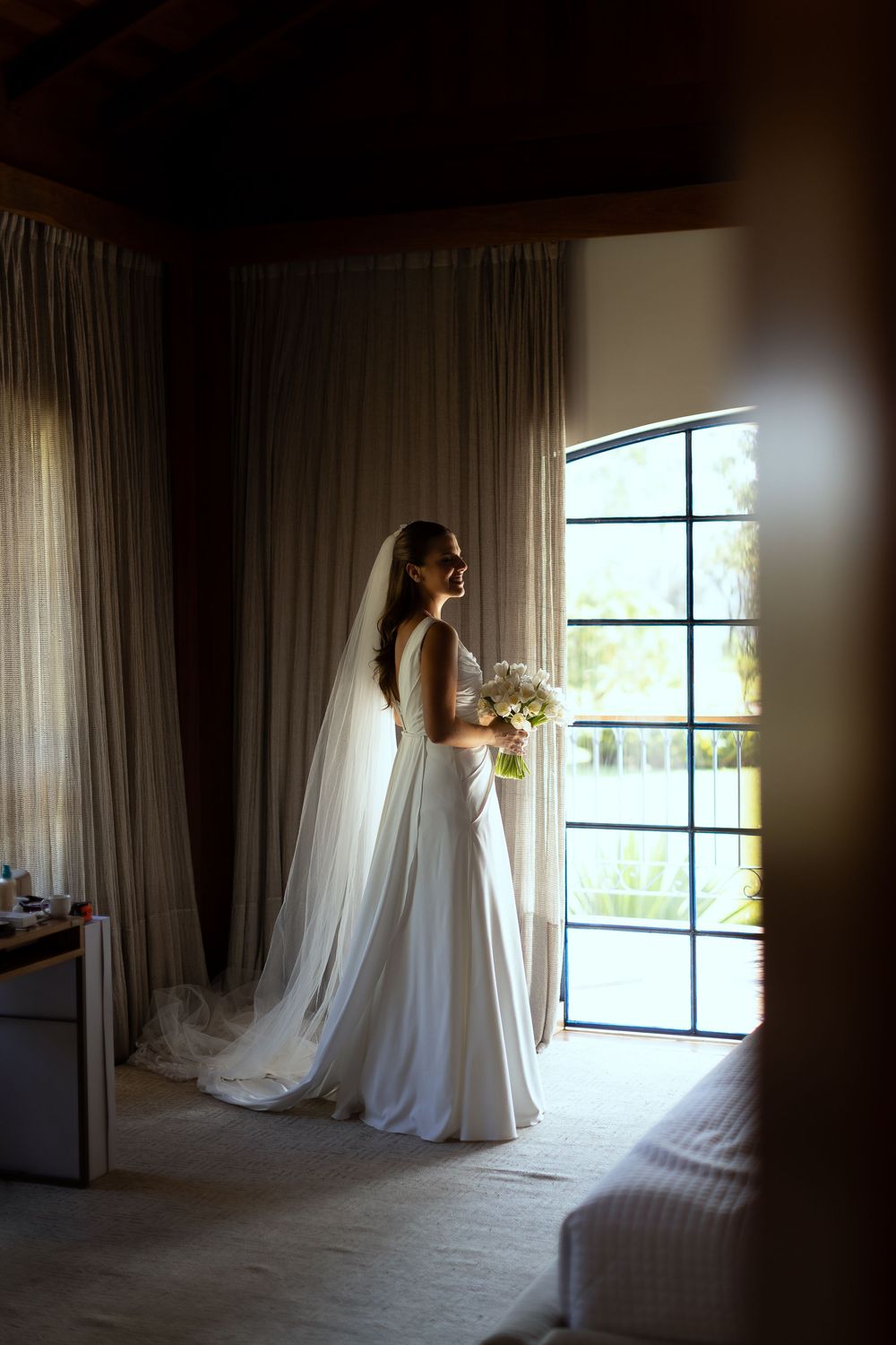 Bride in white gown and veil holds bouquet while gazing out large window in softly lit room.