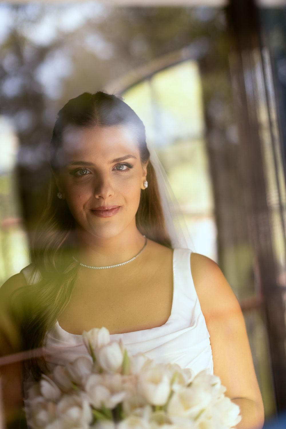 Elegant bride in white dress holding tulip bouquet, photographed through glass window with natural light reflection.