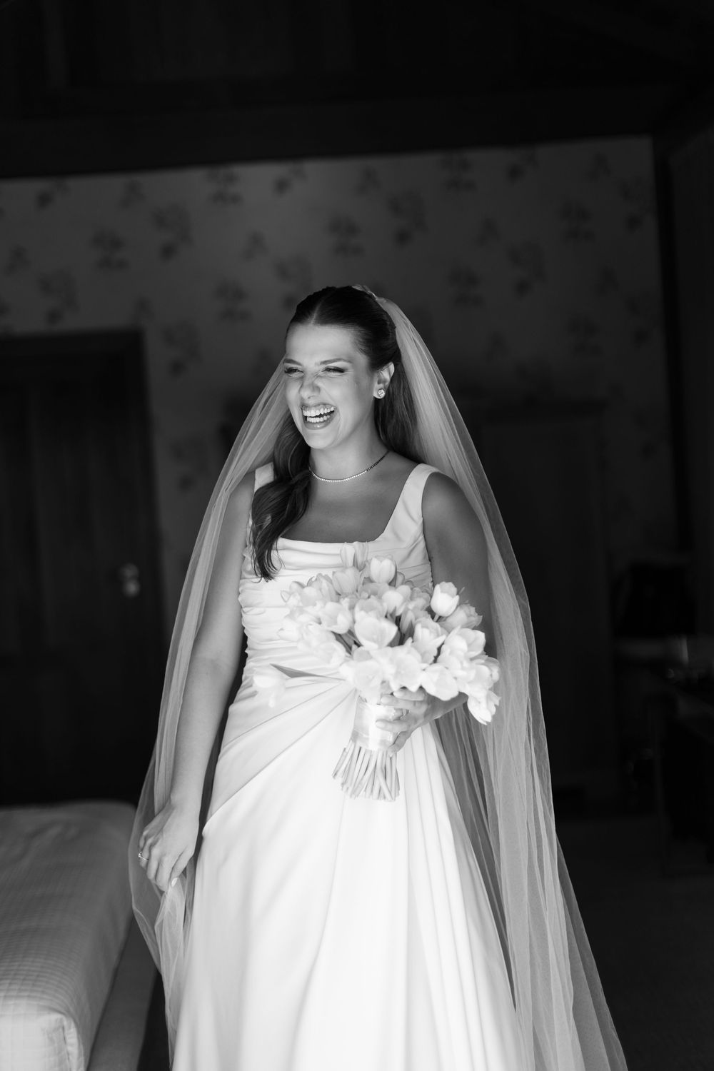 A radiant bride in a white gown and veil holds a bouquet of roses, laughing joyfully in a black and white photo.