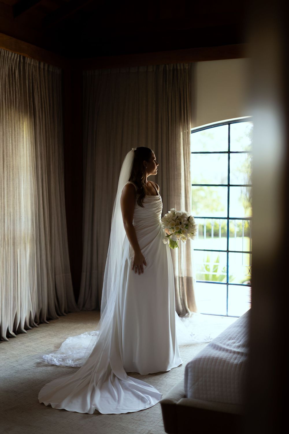 Bride in white gown and veil holding bouquet stands by sunlit window in elegant room.