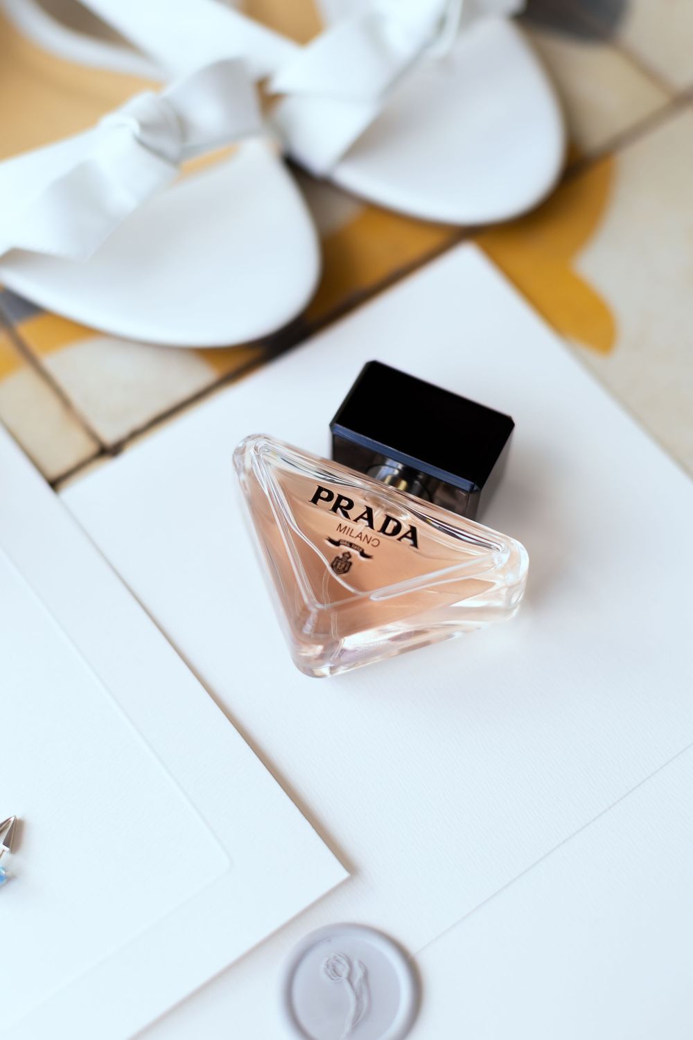 Prada perfume bottle with black cap on white paper, beside white heeled sandals and a wax seal.