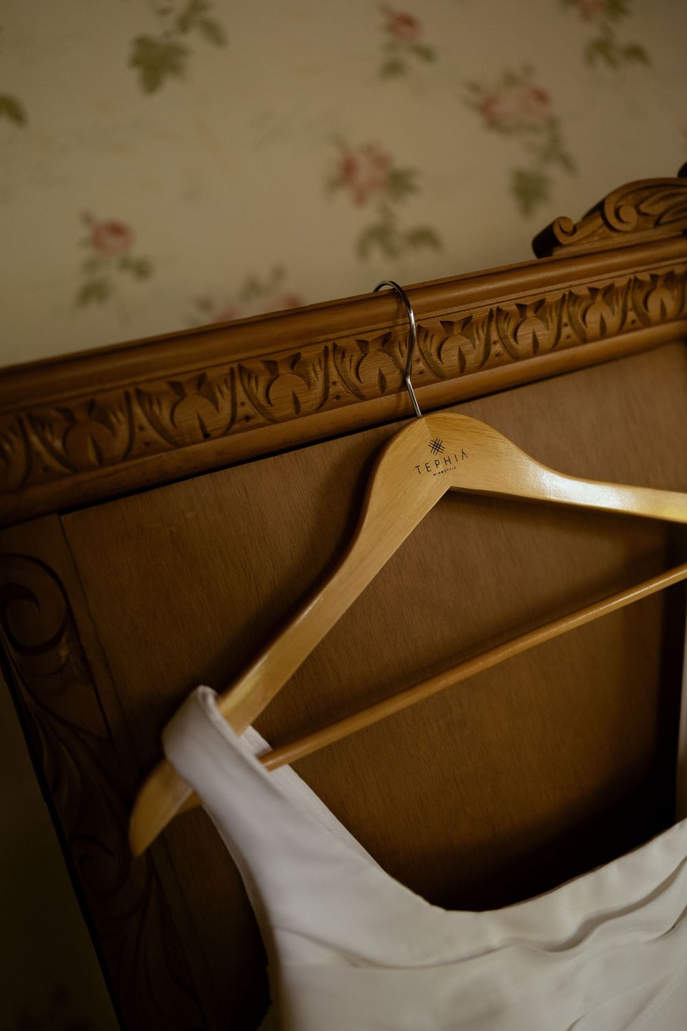 White wedding dress on wooden hanger displayed on ornate headboard with floral wallpaper backdrop.