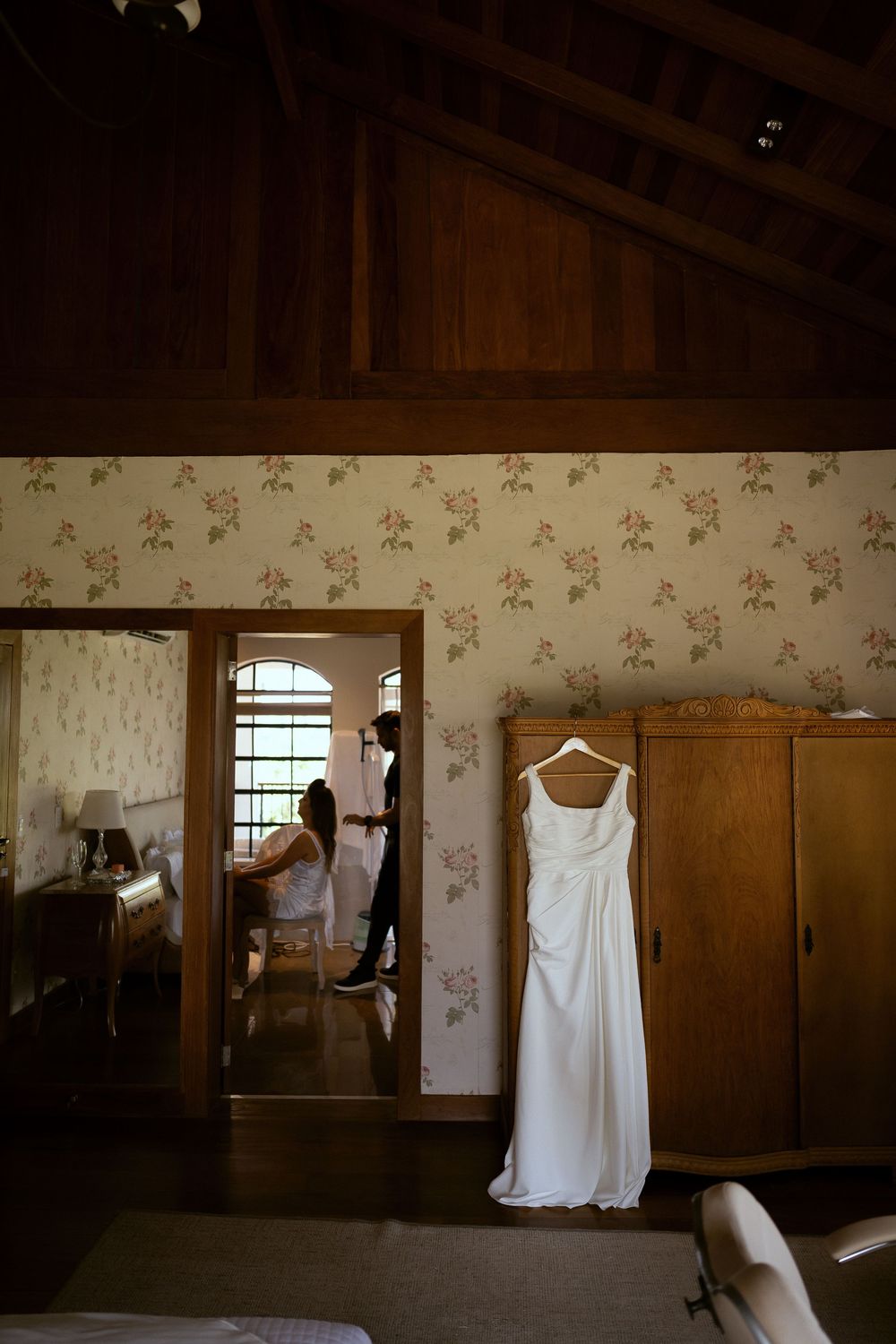 White wedding dress hanging on wooden armoire in floral wallpapered room while bride prepares in background.