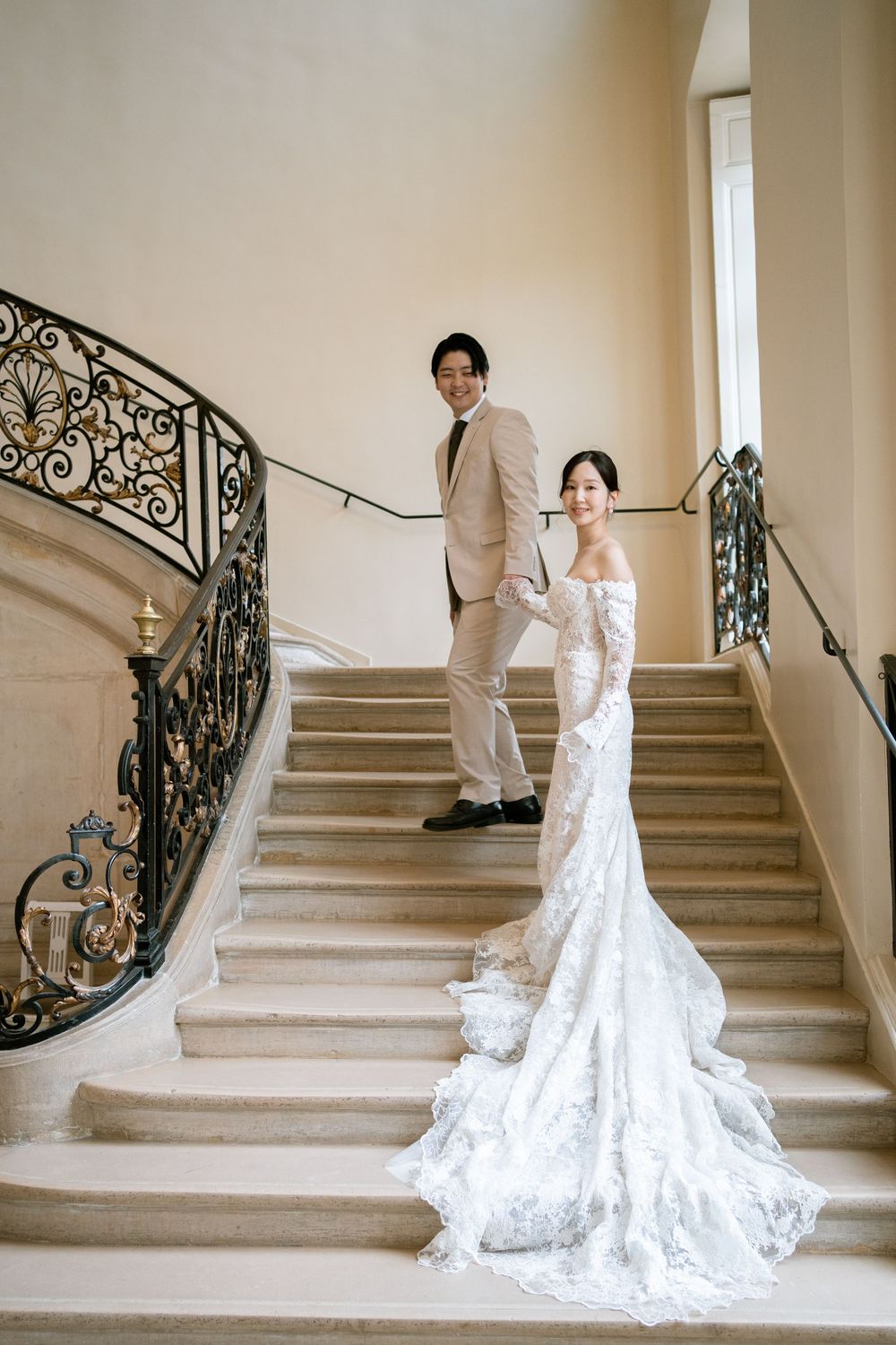 Bride in lace gown and groom in beige suit pose on elegant marble staircase. paris editorial wedding photographer, best paris film photos